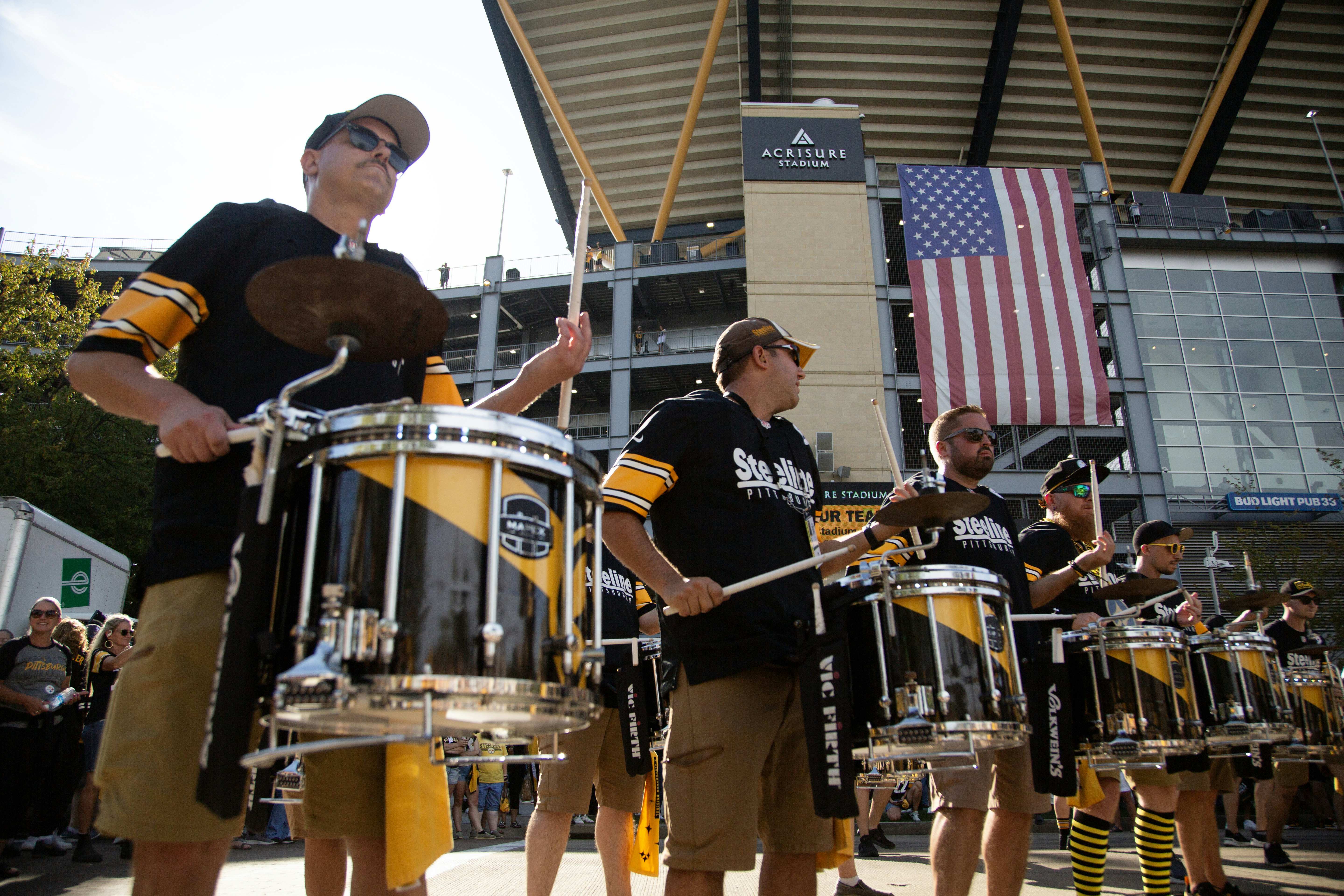 A line of snare drummers play in front of the Pittsburgh Steelers stadium wearing black and gold Steelers jerseys.