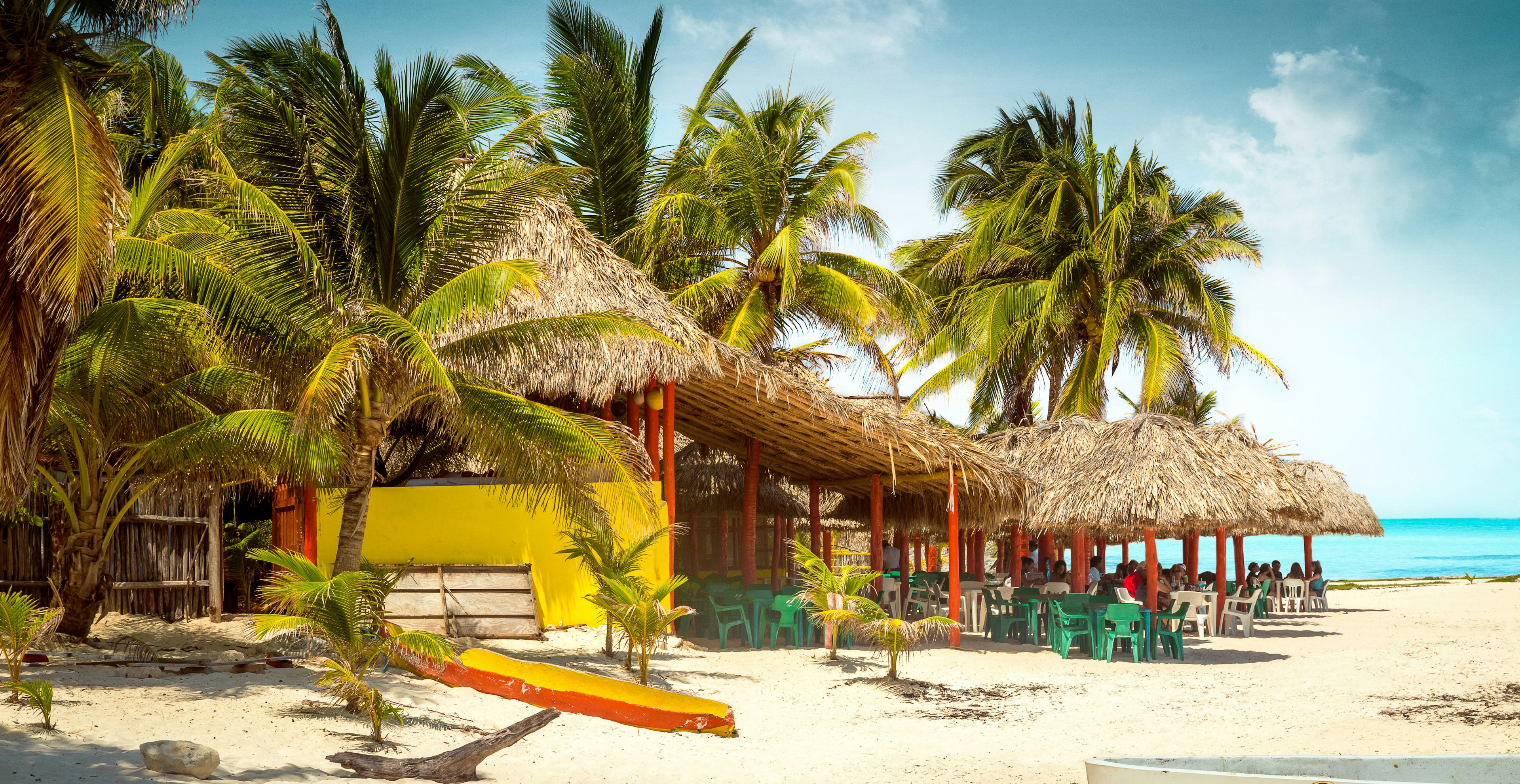 A thatched-roofed tropical bar sits beneath hulking palms on a beach at Cozumel island. The bar is painted bright yellow and red with green and white plastic chairs. It's partly obscured by green palm trees and sits on a white-sand beach with a jewel-blue sea in the background.