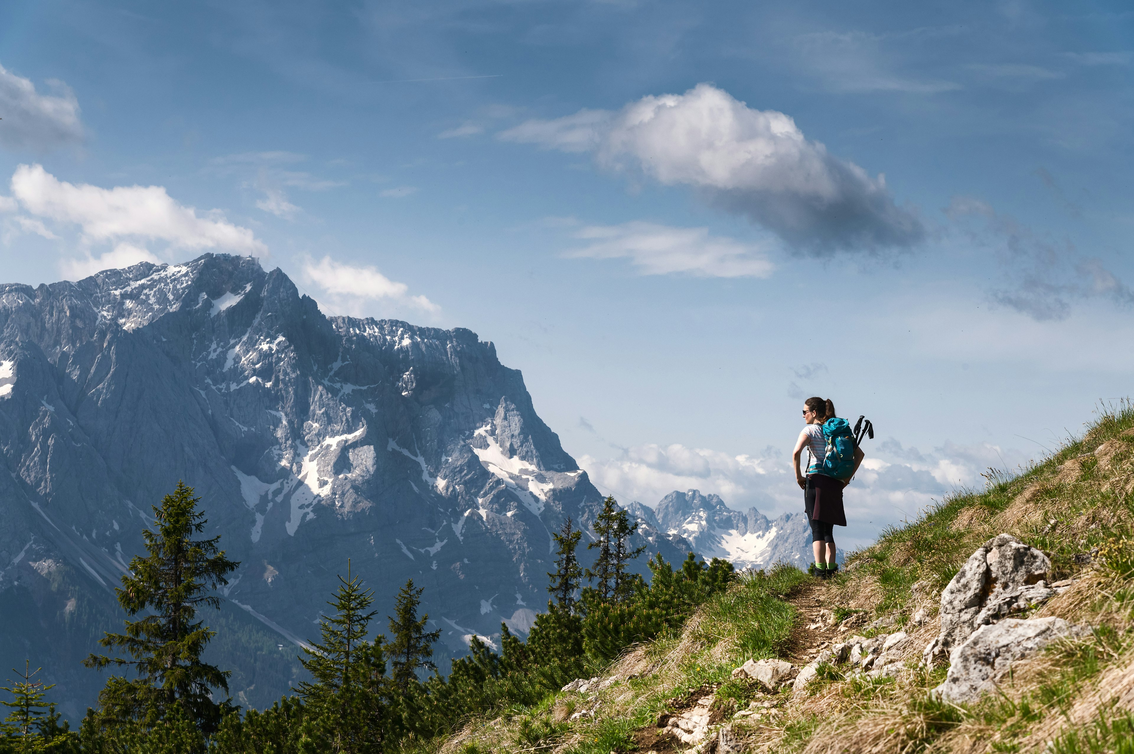 A female hiker with a backpack and climbing poles stands on a scrub-covered hillside looking out on the snow-capped peaks of the German Alps, including the Zugspitze, Germany's highest peak.