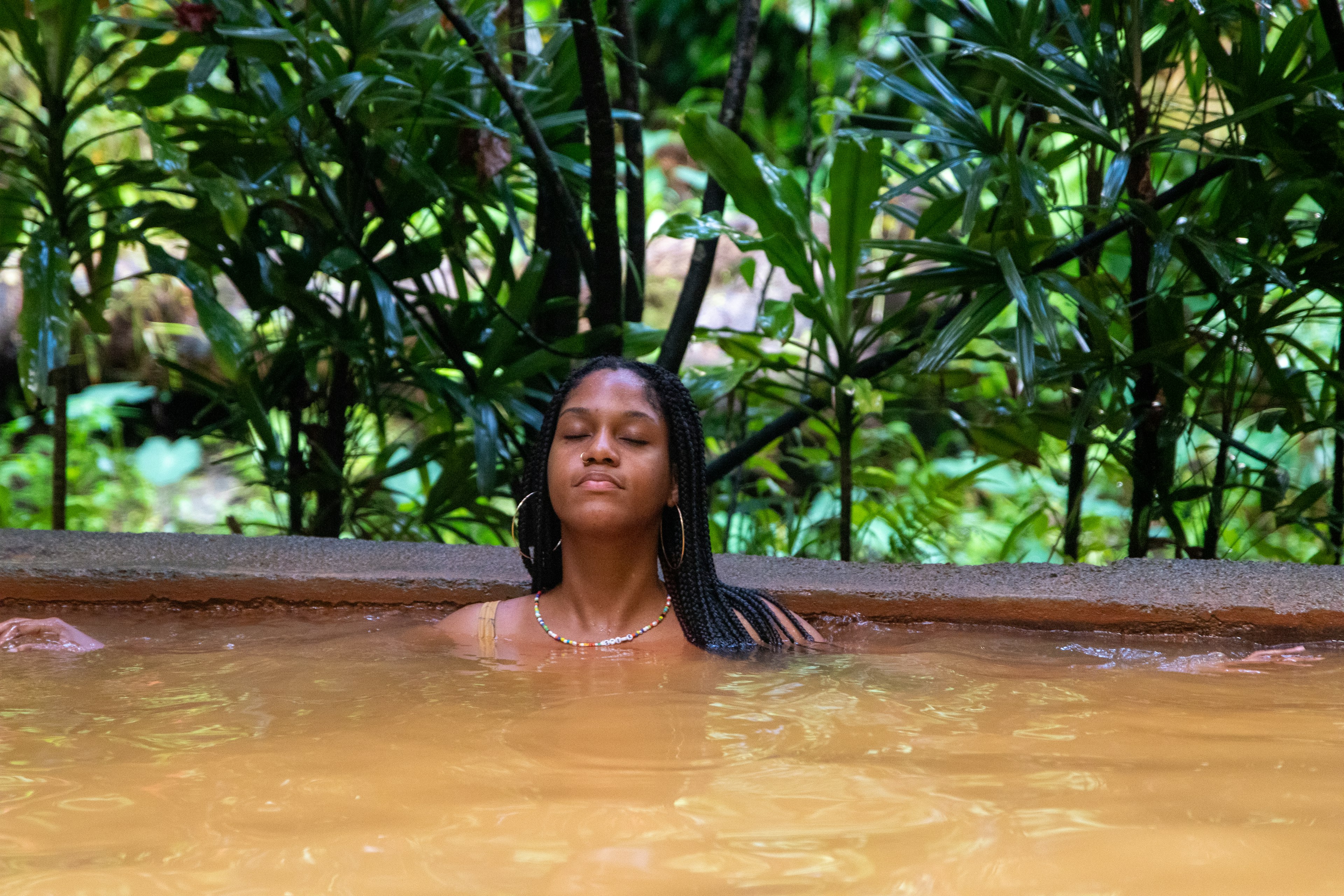 A woman with braids, hoop earrings and a beaded necklace closes her eyes as she sits in murky thermal waters up to her neck Tikwen Choglo natural spa, Dominica