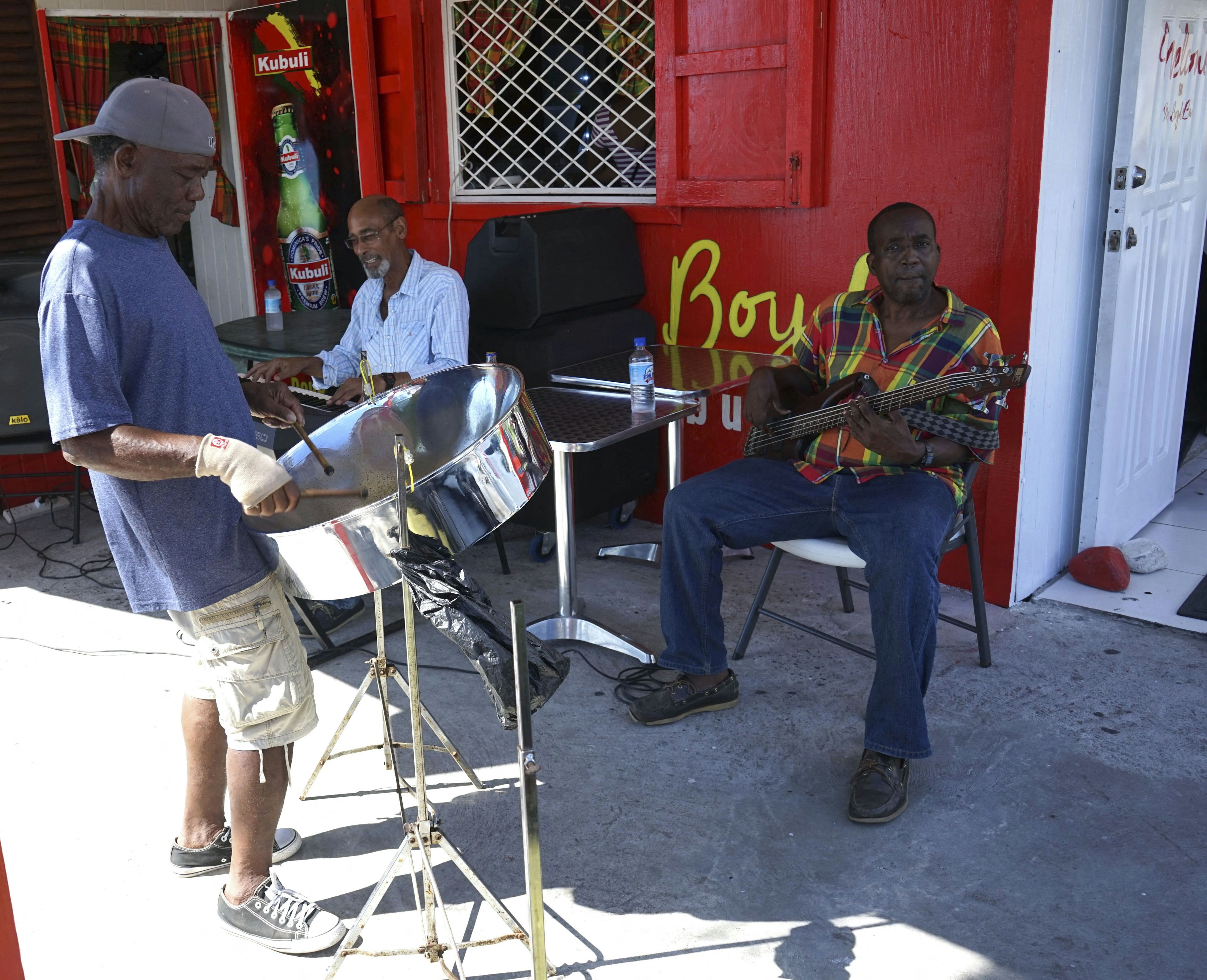 Three musicians – on bass, keyboard and steel drum – perform in the street against a red-painted wall, Roseau, Dominica