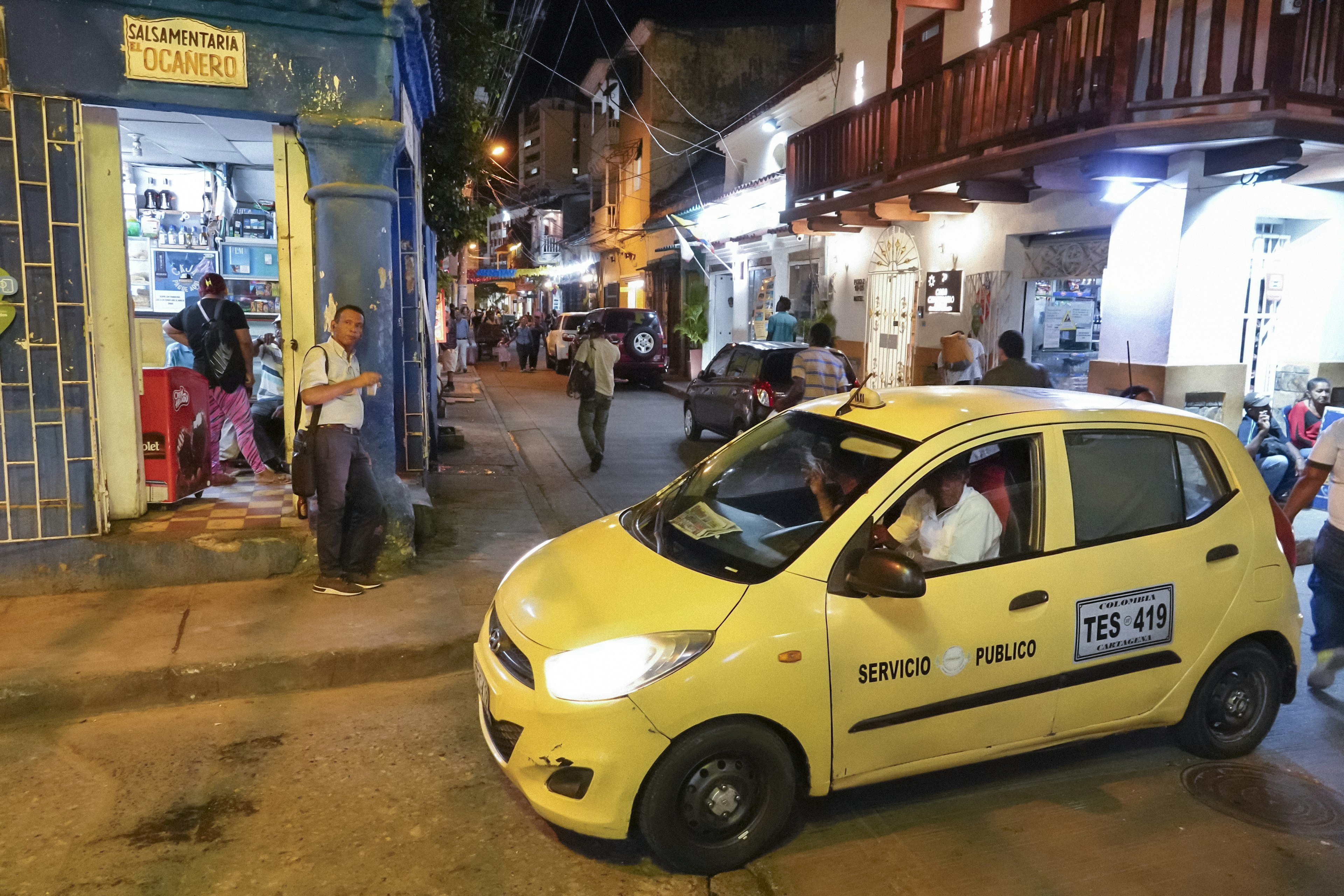 A yellow taxi waits on a curb at night, by a busy street with shops and illuminated façades, Getsemaní, Cartagena, Colombia