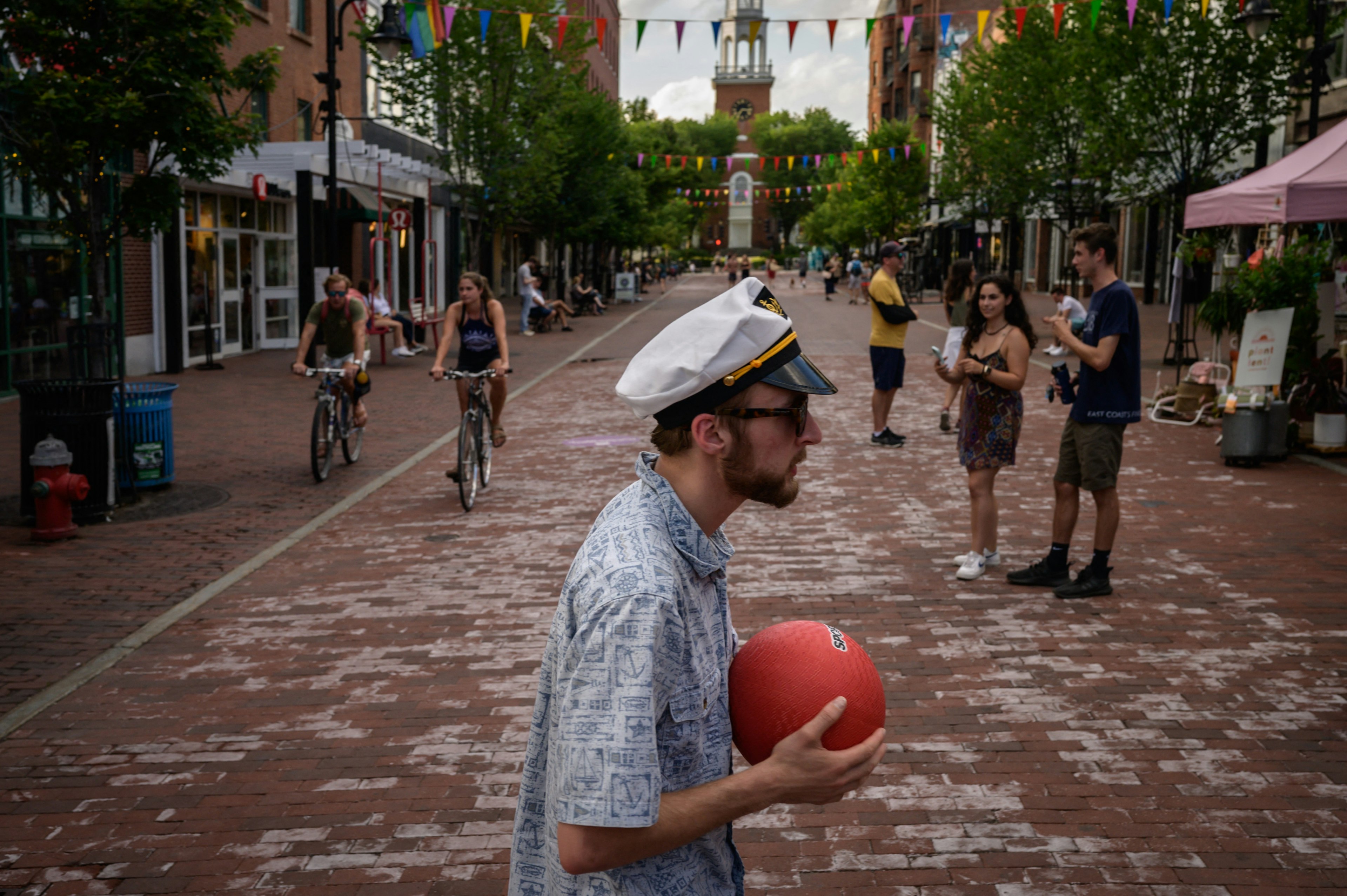 A man in a sailors hat holding a red ball is pictured along with other people walking and cycling on Church St in downtown Burlington, Vermont, New England, USA