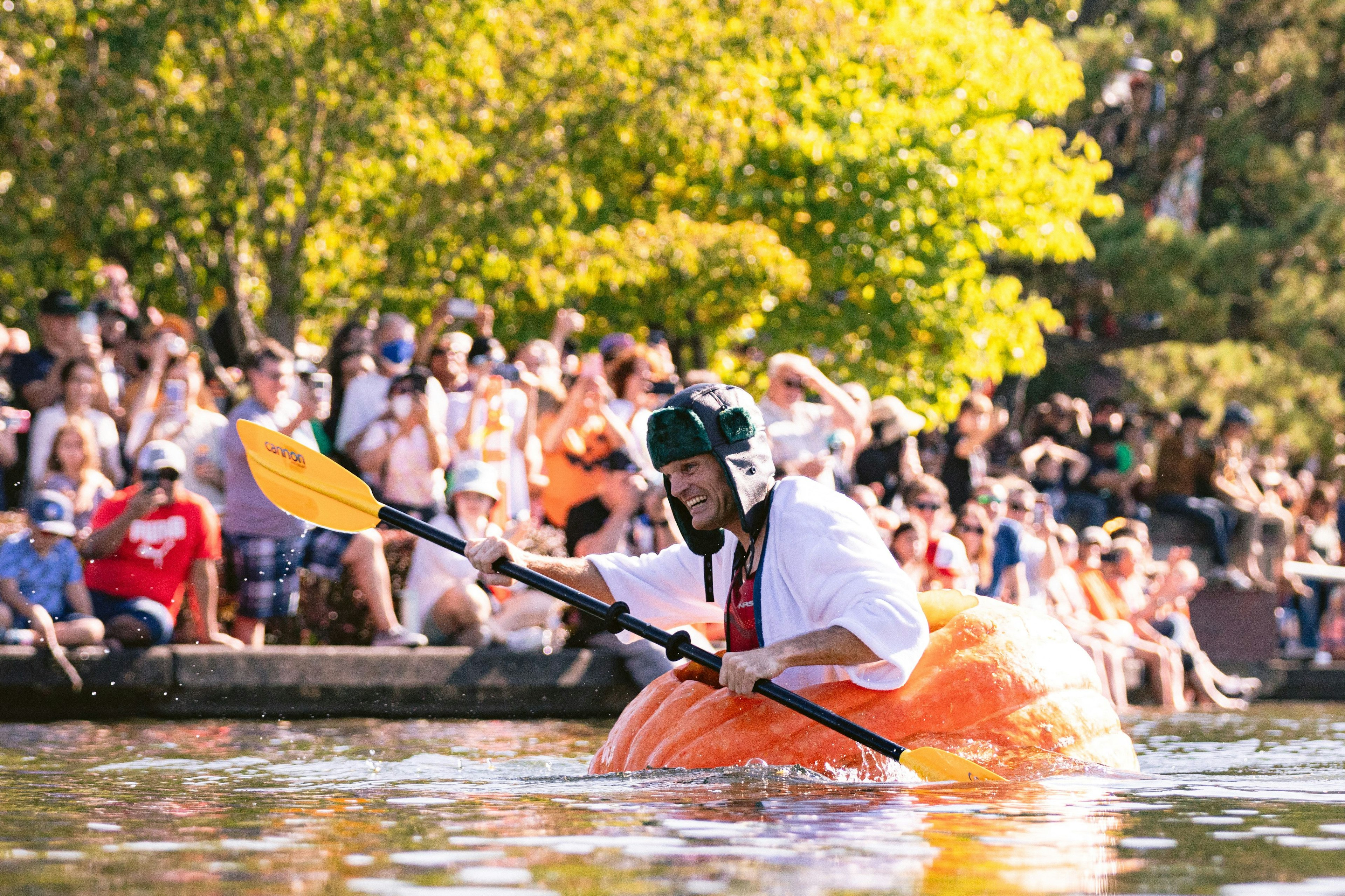 A man in a winter hat paddles in a giant pumpkin in water as crowds look on on a sunny fall day