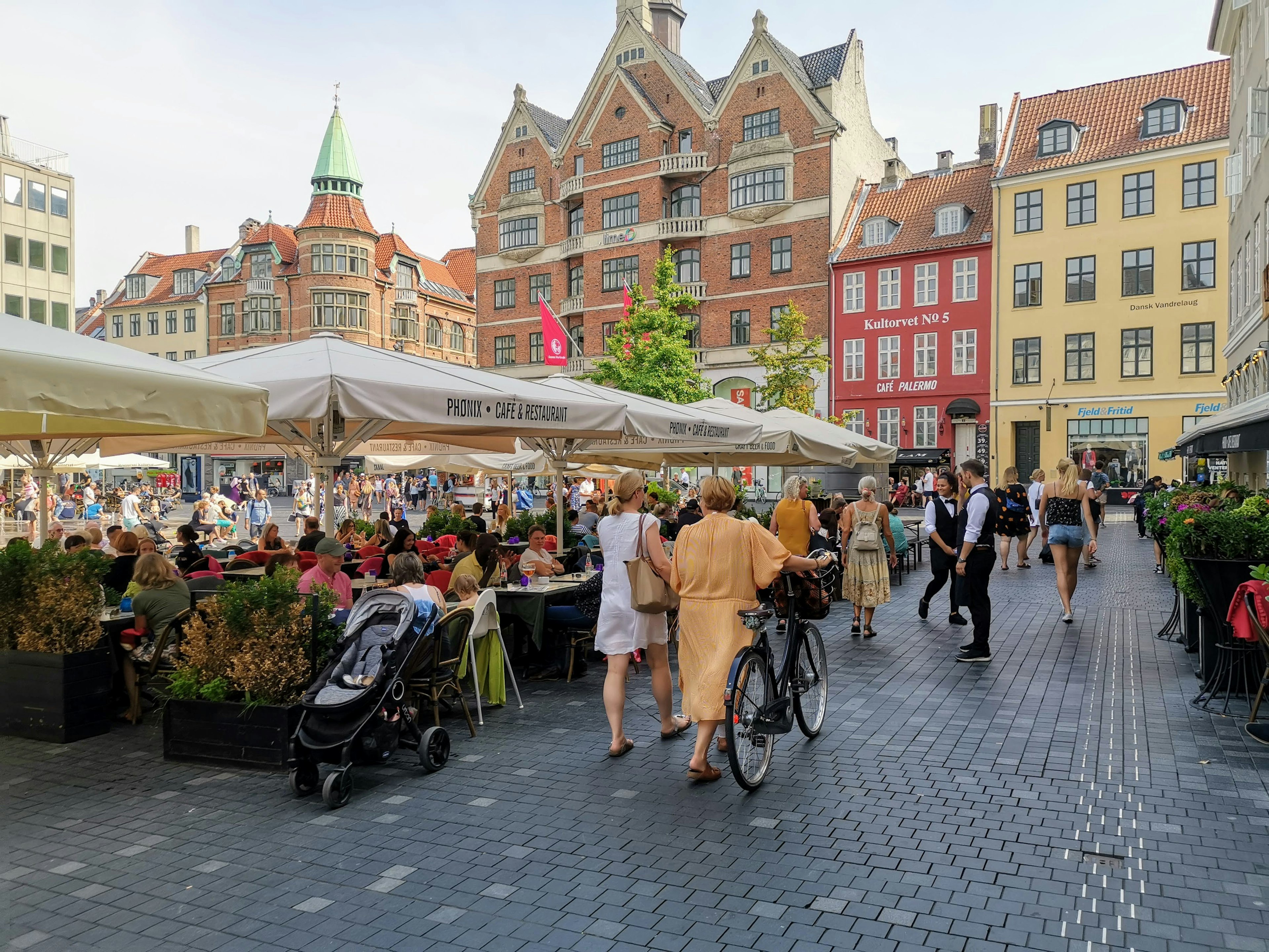 People sat at tables outside a restaurant in a city square as people walk by wheeling a bike