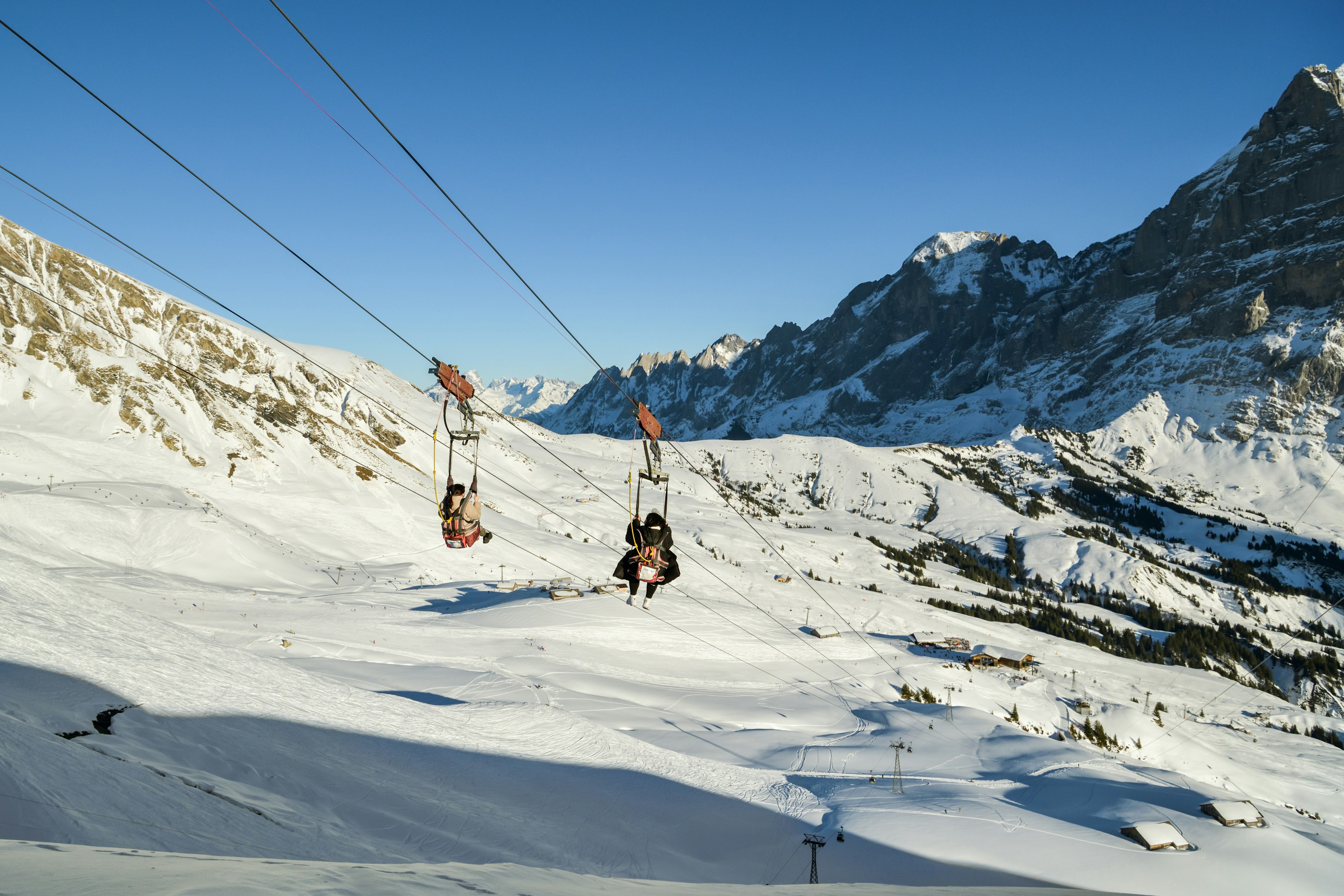 Two people on a zip line fly downwards surrounded by a snowy mountain landscape