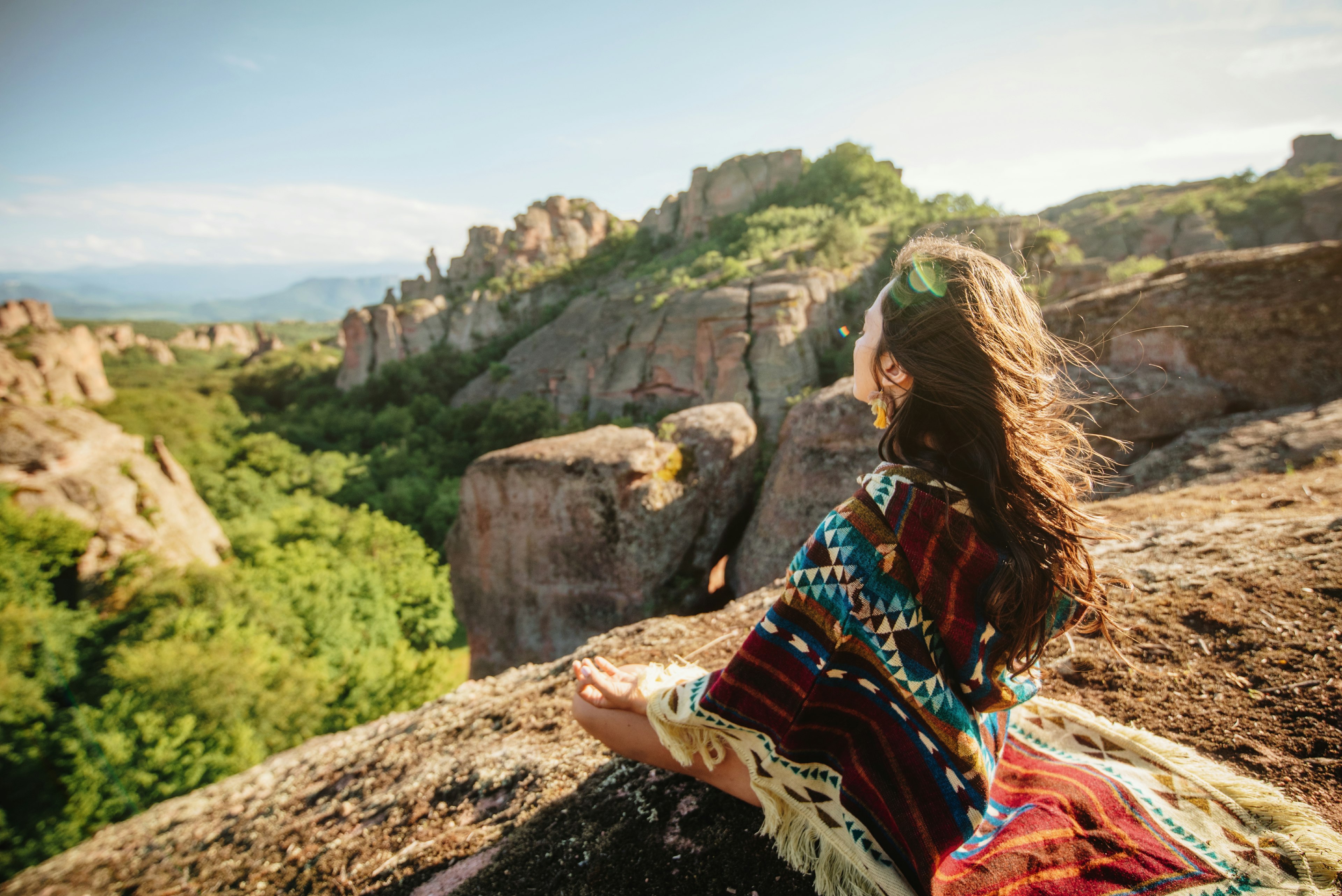 Young boho woman wearing a geometric serape meditating on the edge of a cliff overlooking greenery near Sedona, Arizona