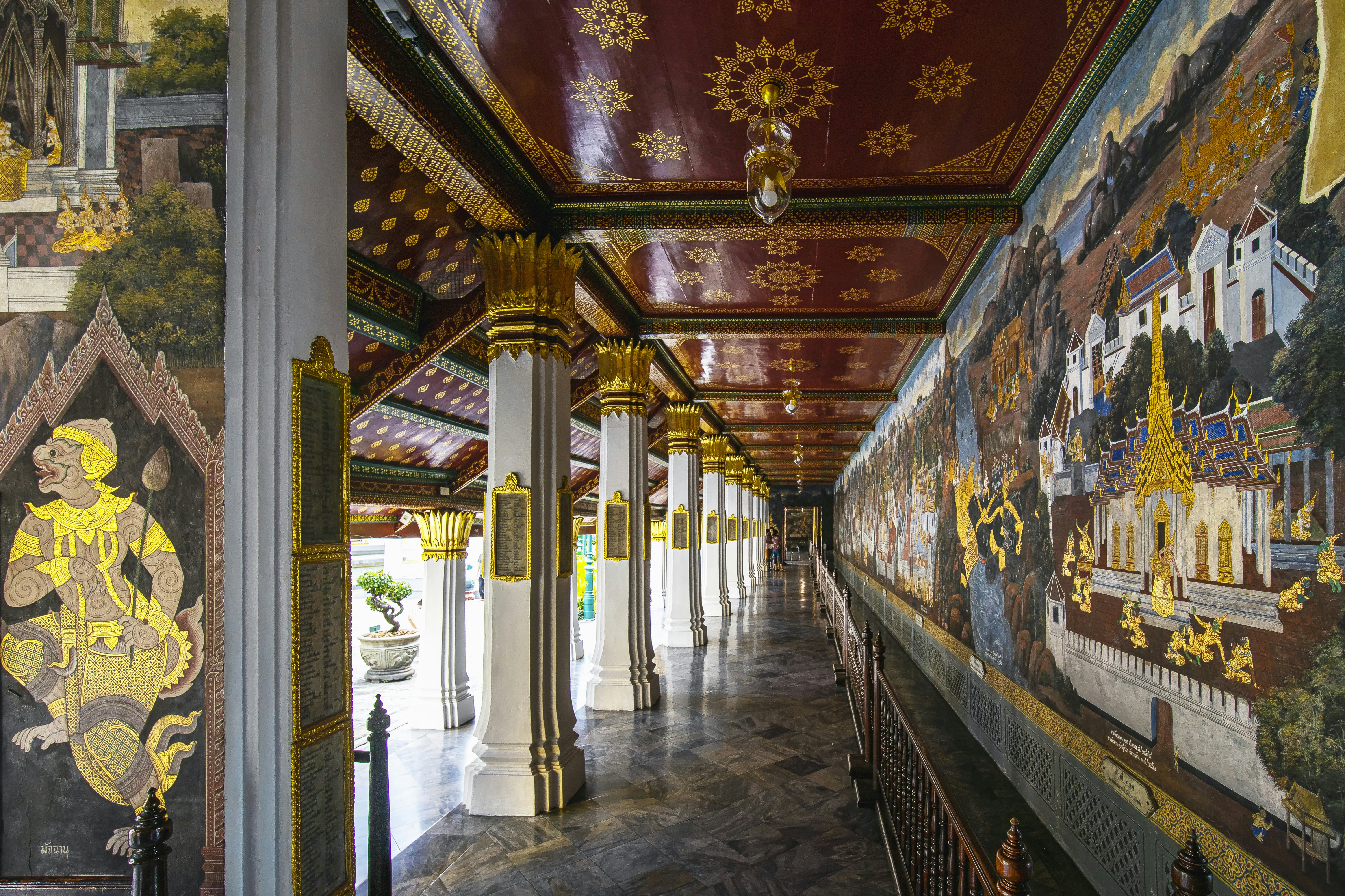 A hallway surrounding an open courtyard with elaborate, gilded pillars on one side and the brightly colored and gilded Ramakian Murals at Wat Phra Kaew temple, Bangkok