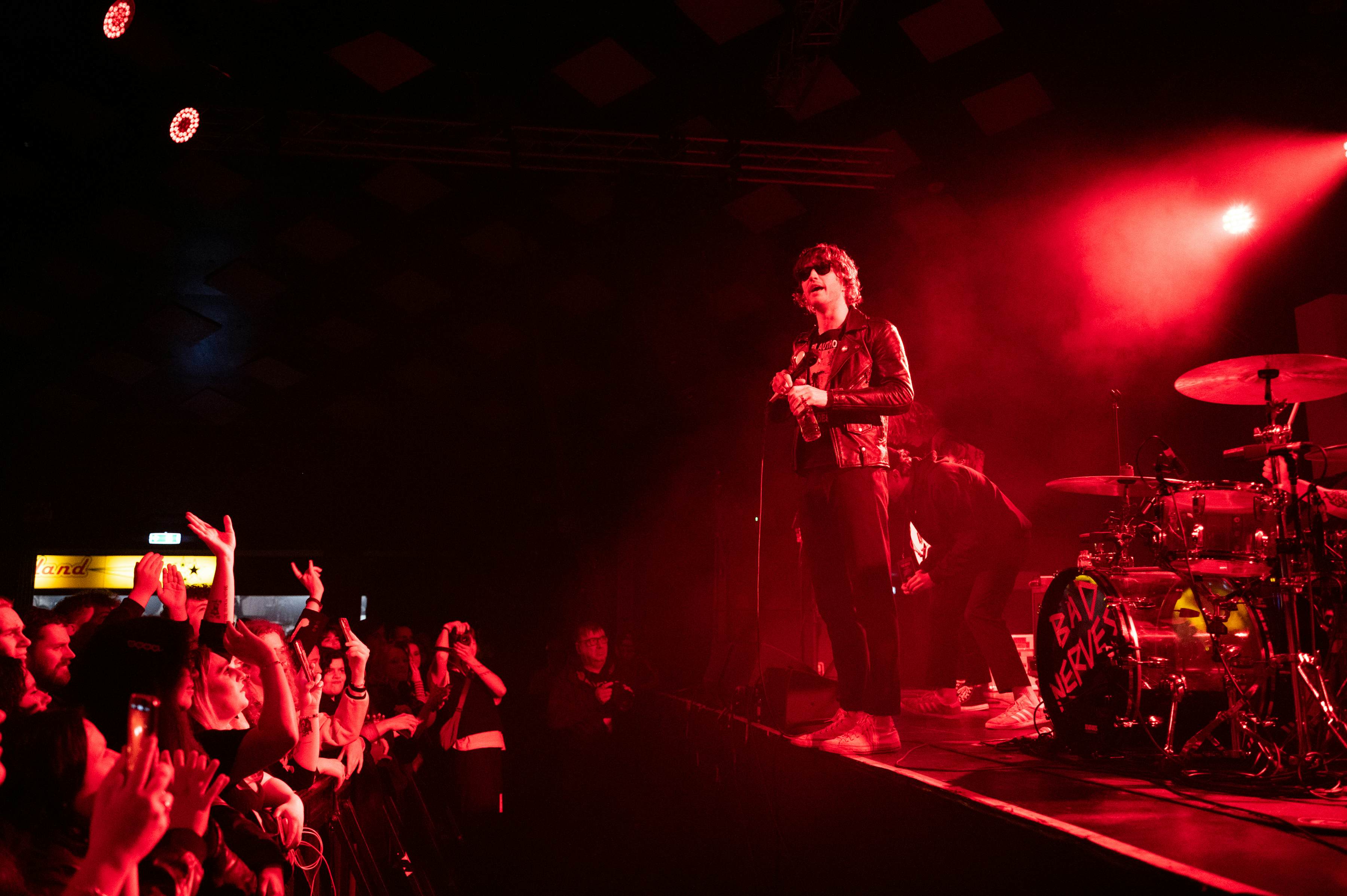 A rock performer in sunglasses and a leather jacket performing on stage, bathed in red light, as fans look on, Barrowlands, Glasgow, Scotland, UK