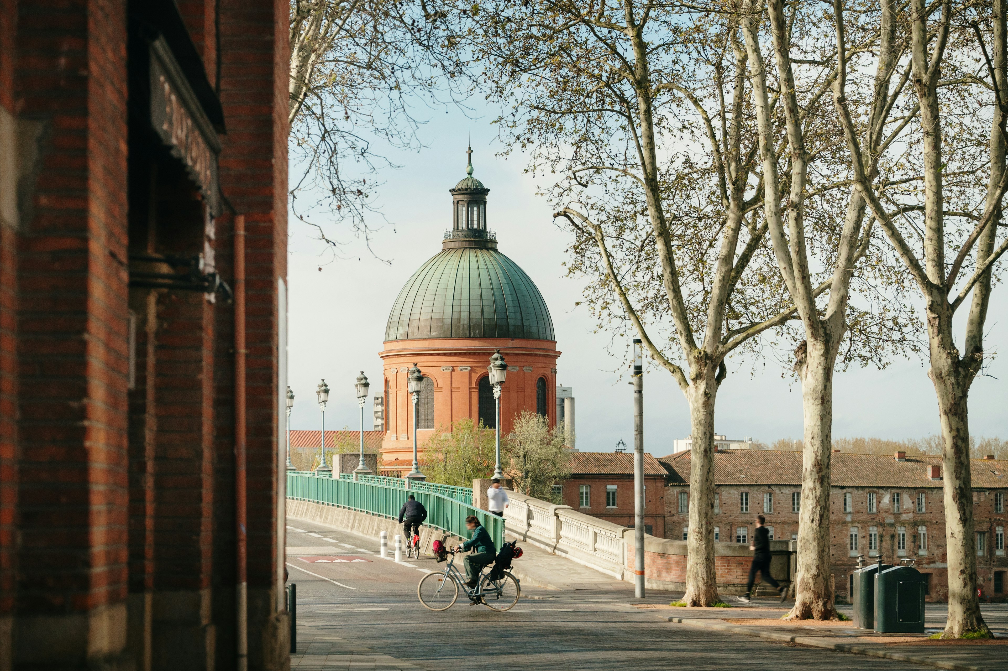 A person bicycles along a riverfront path in Toulouse, France, with the Dôme de la Grave visible in the distance