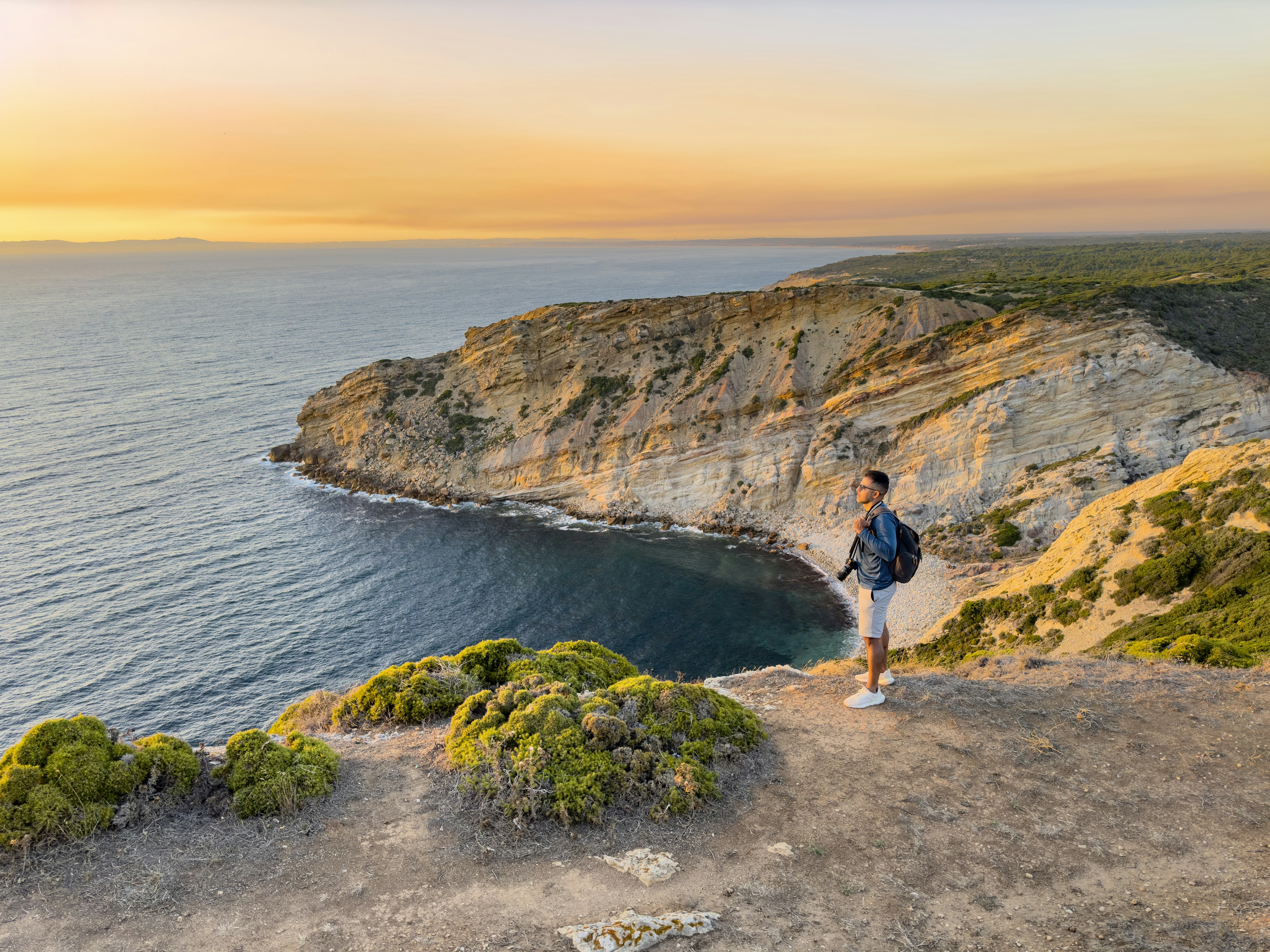 A tourist stands holding a camera gazing out at sunset from a cliff above a beach