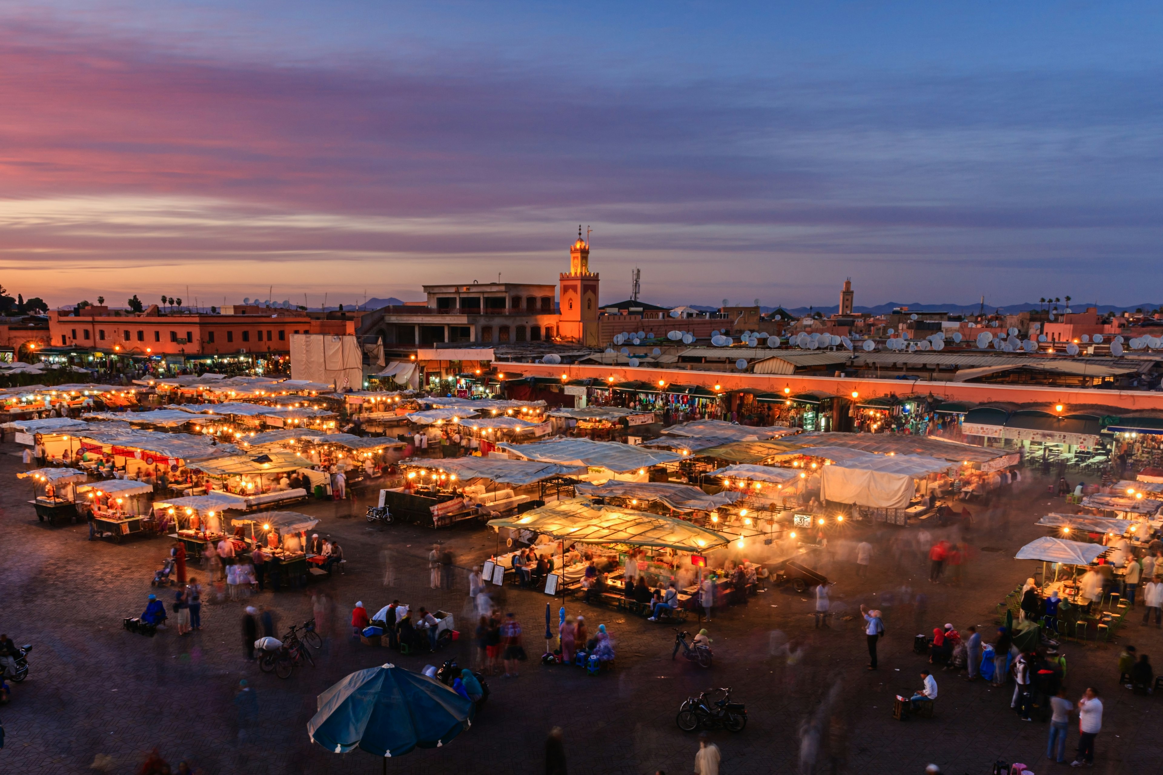 Aerial of a crowd at Djemaa El Fna square during the early evening in Marrakesh's Medina quarter; moving people are blurred between the umbrella-covered food stands. The mosque's minaret stands tall against the violet sky in the background.