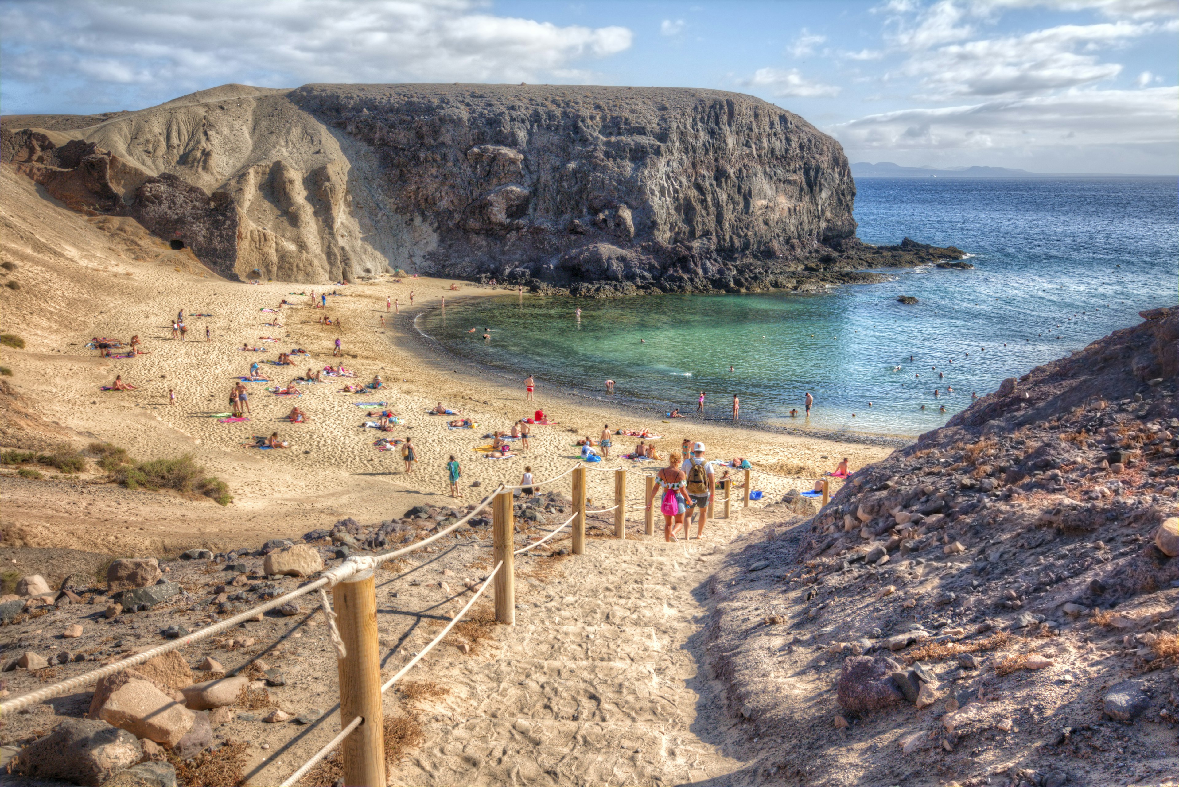 People descend steps to small beach with blue waters, in between jagged rock formations