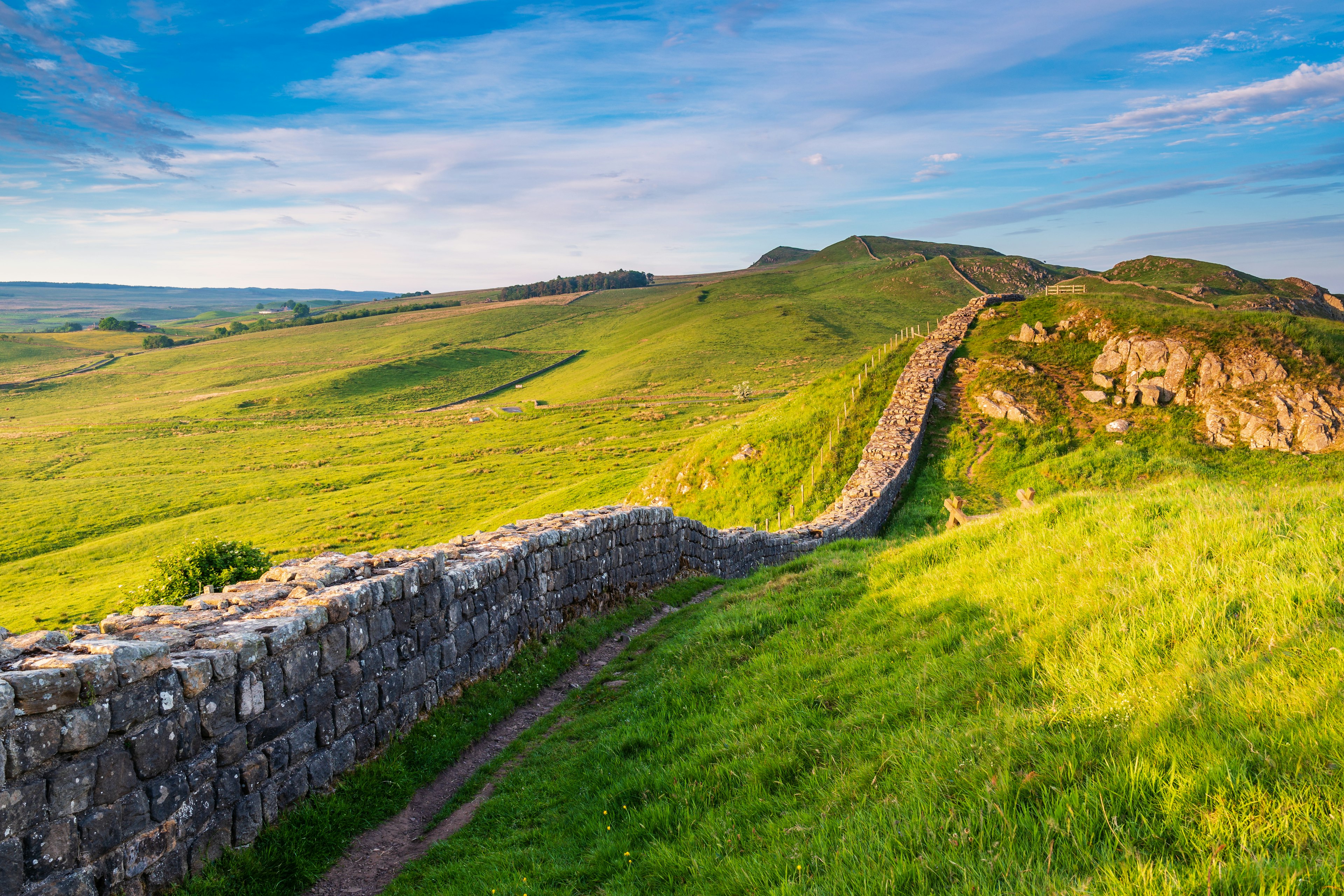 A dry stone wall running through hilly green countryside with cloudy blue skies.