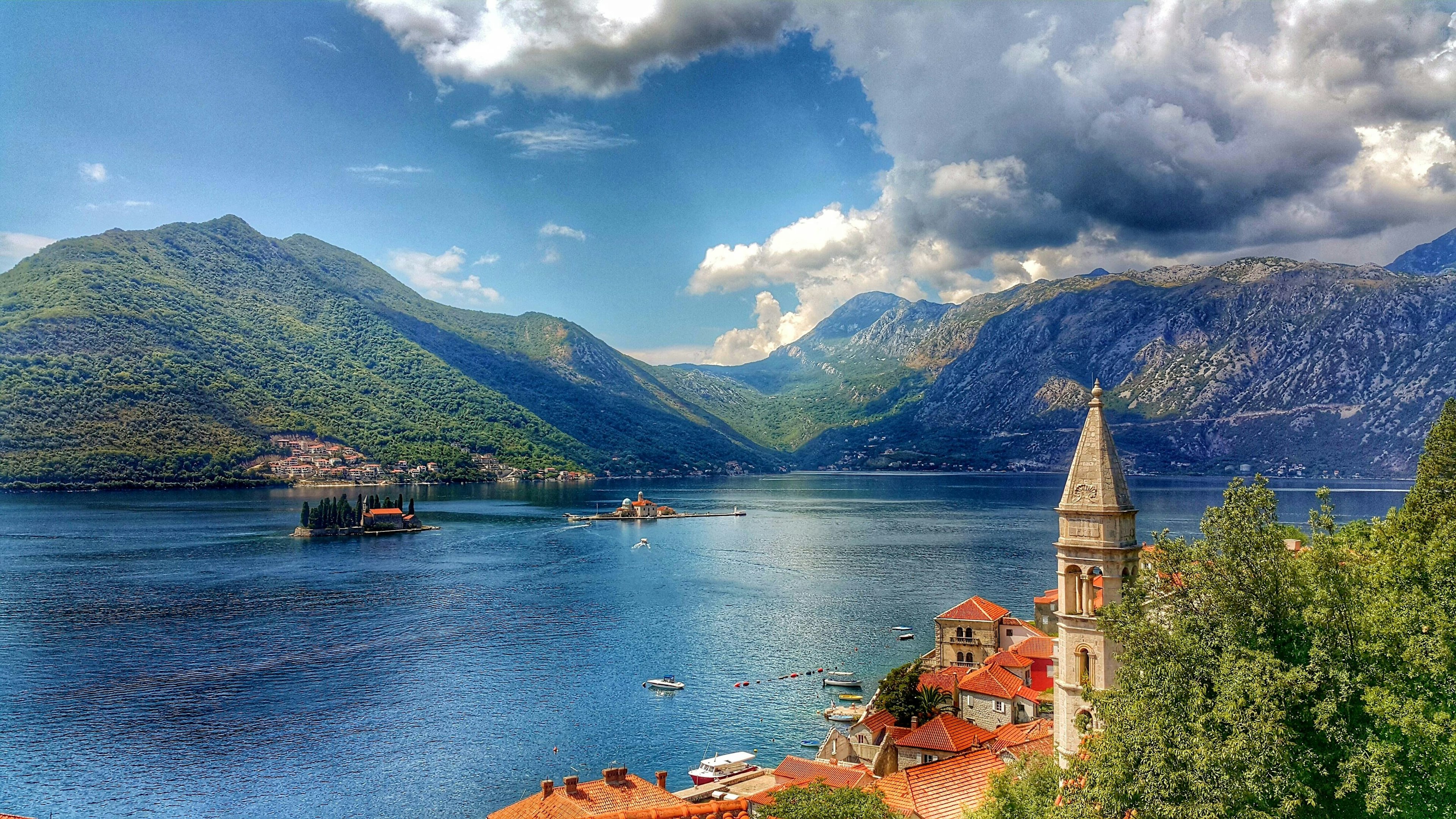 A red-roofed town sits at the edge of a bay of water, with hills in the background. Dramatic clouds cast shadows over the hillsides