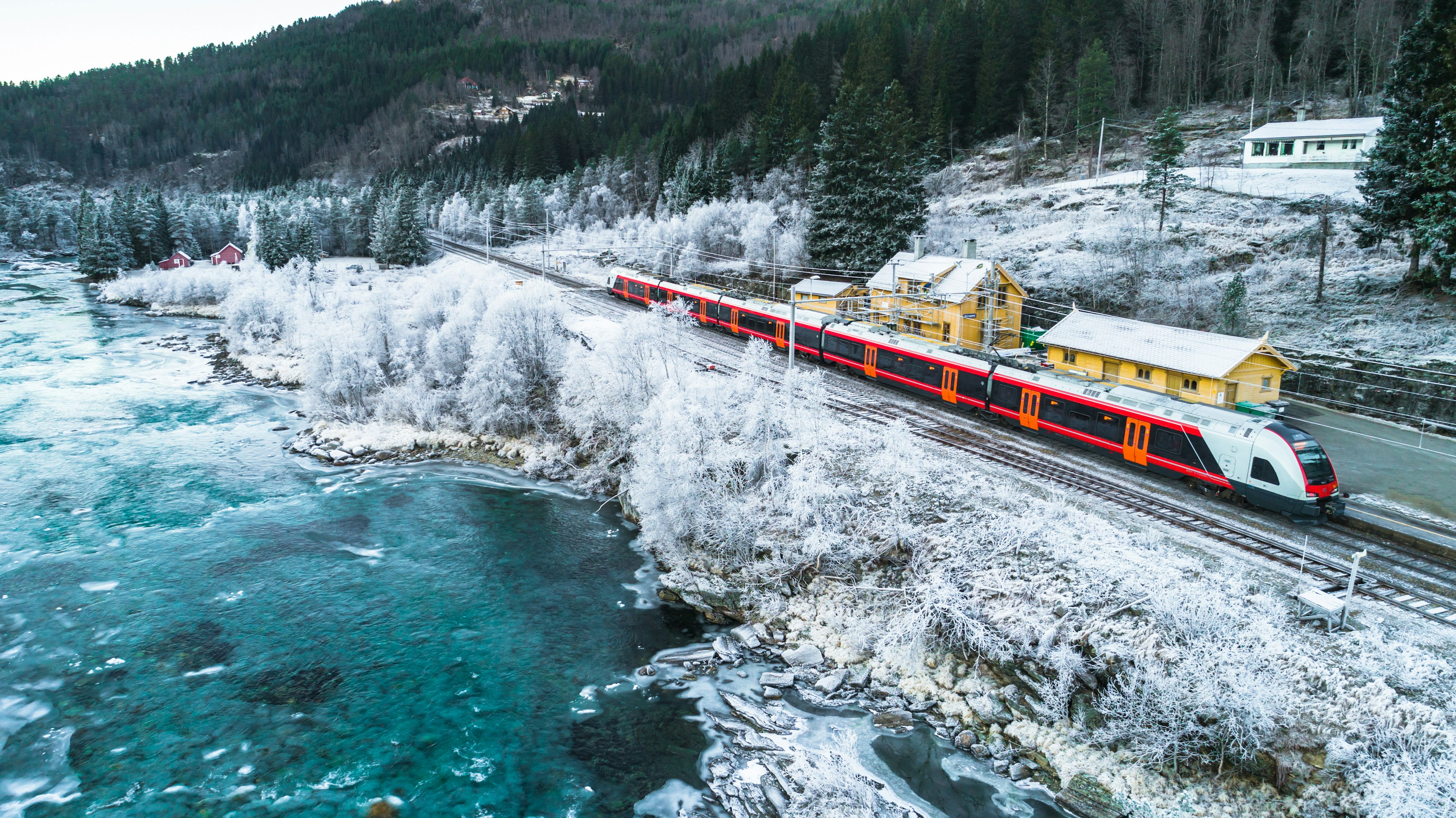 A red train travels through snowy surrounds, with yellow buildings on the right and a frigid lake to the left of frame.