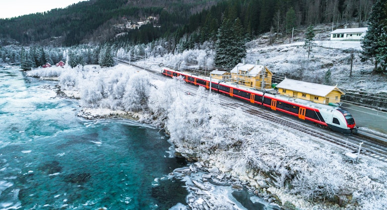 A red train travels through snowy surrounds, with yellow buildings on the right and a frigid lake to the left of frame.