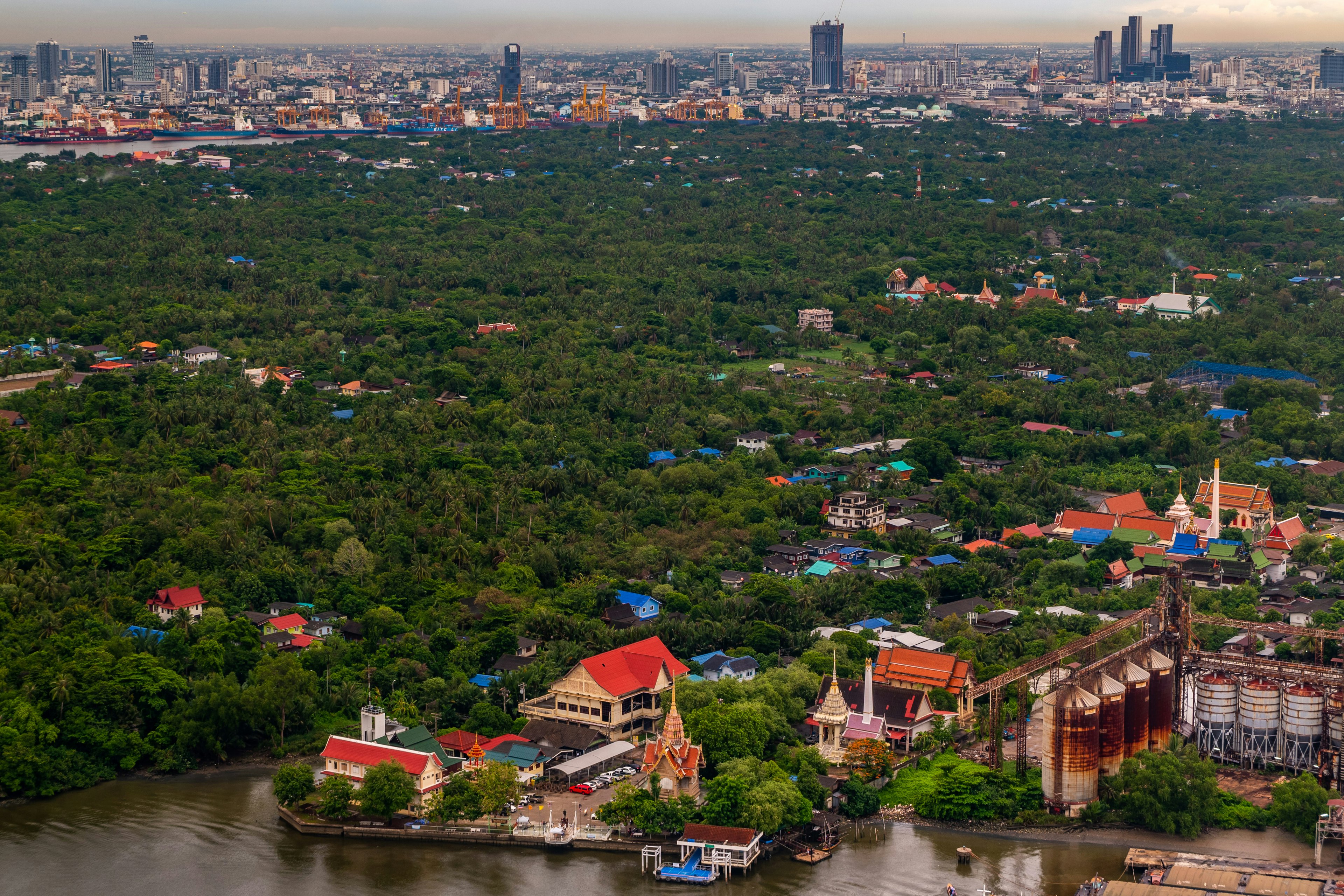 A large green area dense with trees, with a few temples and other buildings standing out. Taller buildings and signs of industry can be seen along the nearby river and the city in the distance