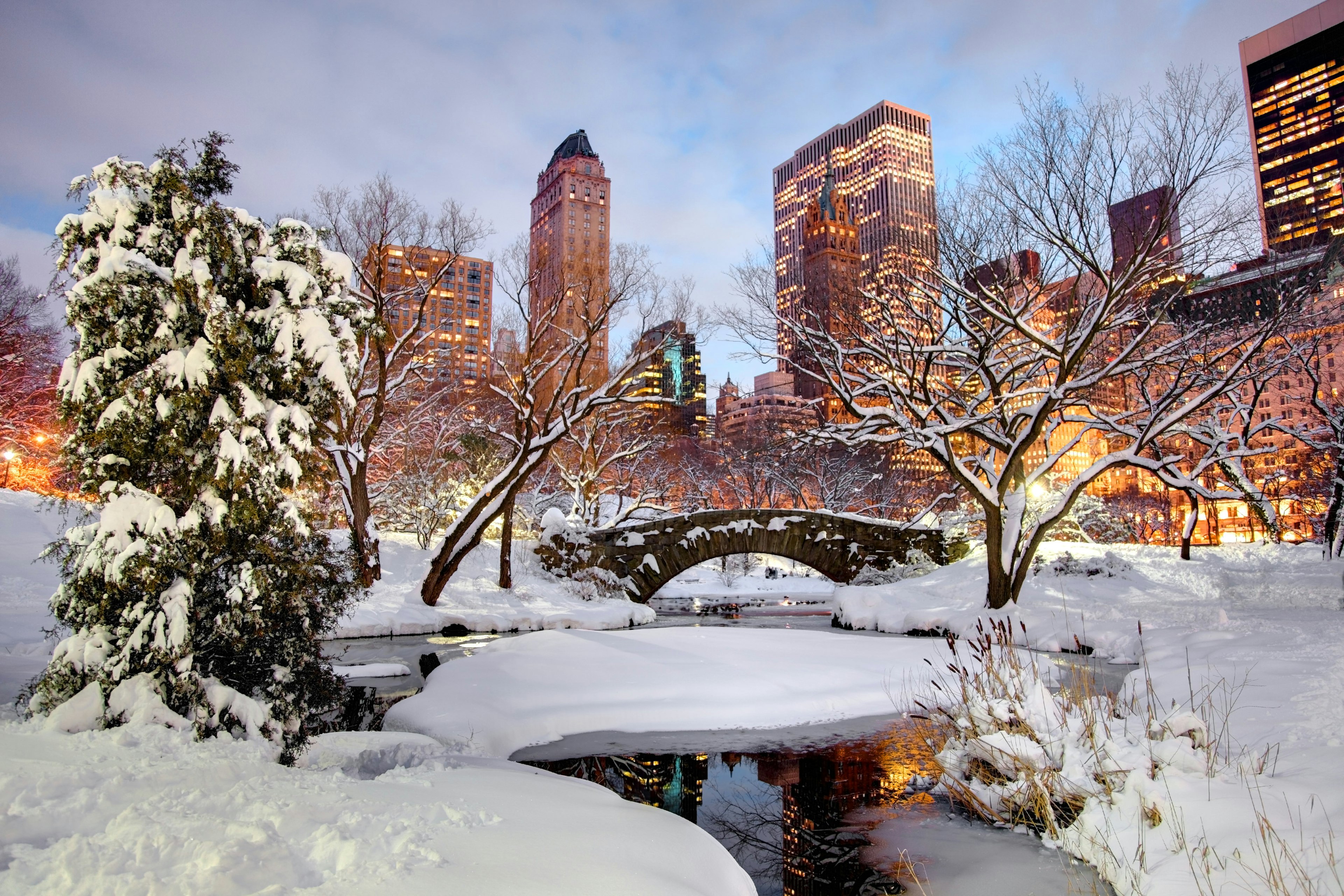 Snow-covered ground and an icey waterway in a city park with a stone bridge over a stream