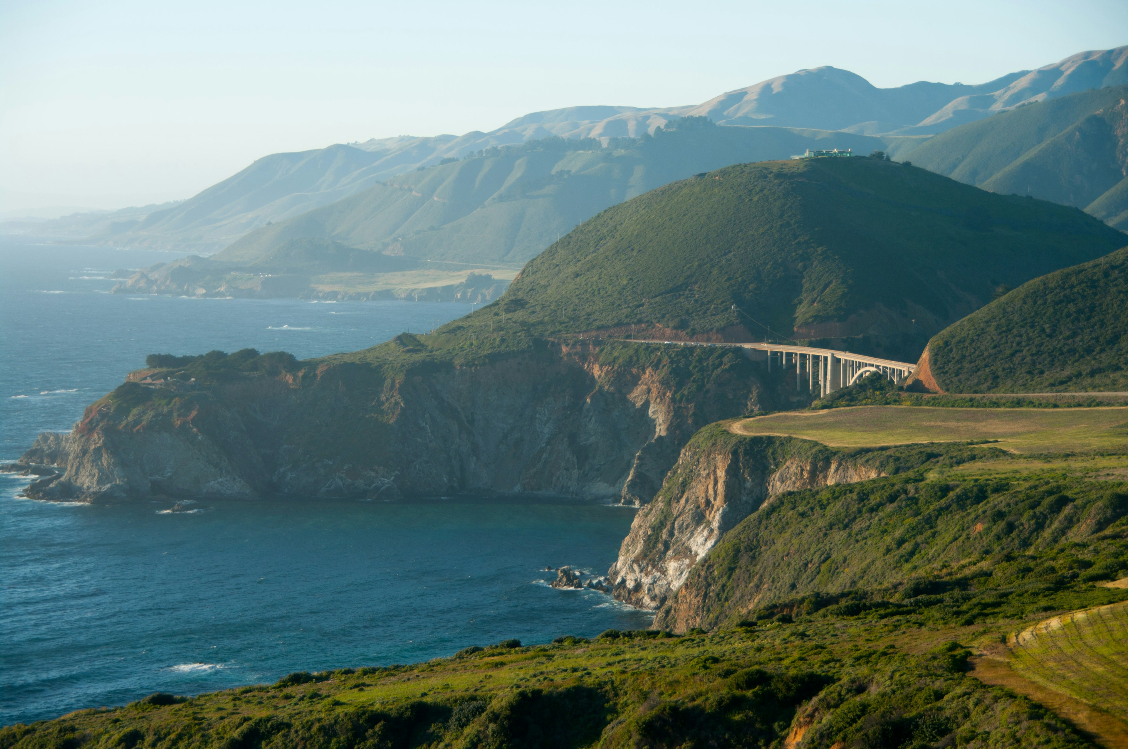 Rolling hills end in cliffs that border the ocean, a single span bridge stretches across the hills.