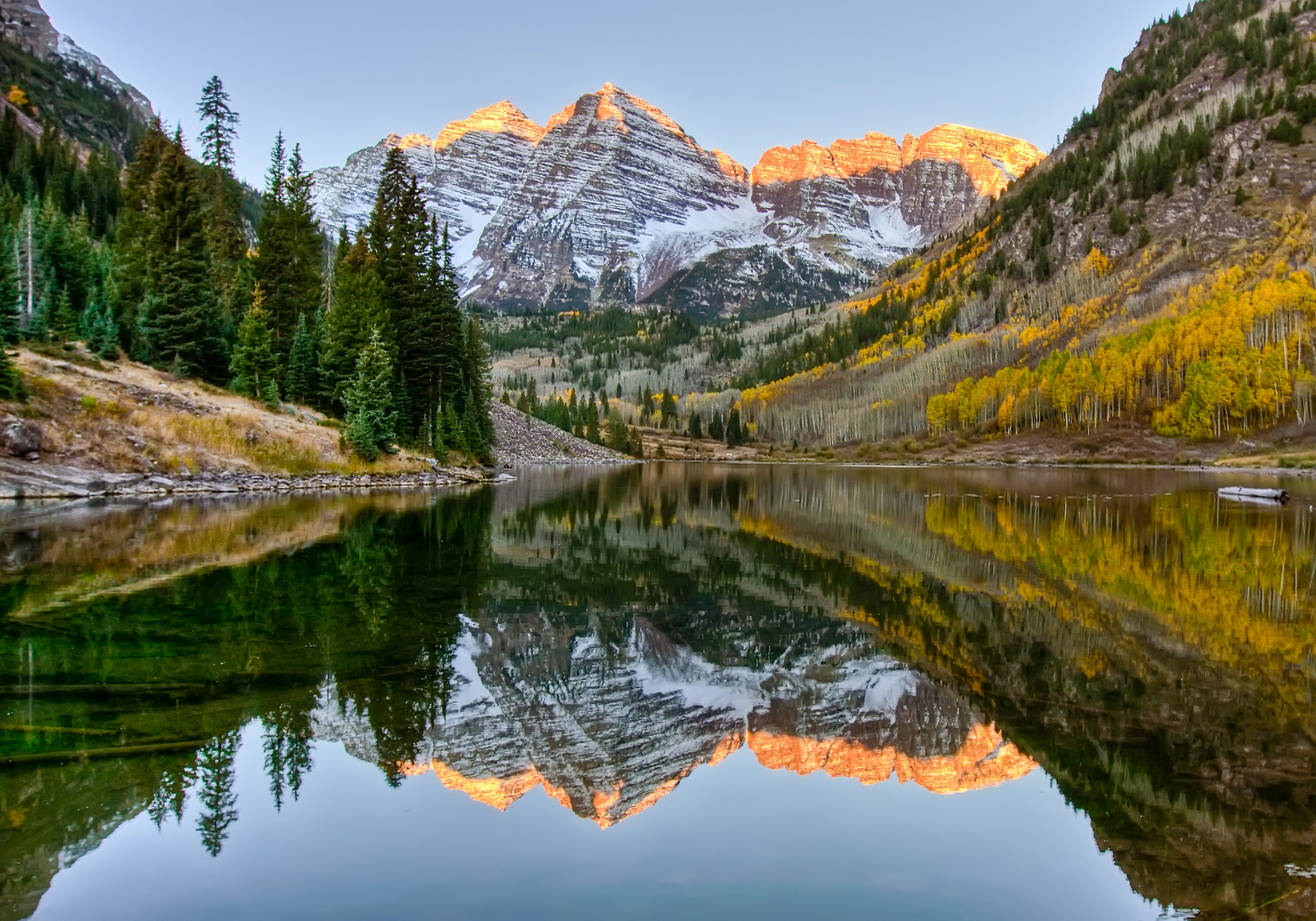 Sunrise hits snow dusted peaks of Maroon Bells while being reflected in a lake below, along with Aspen trees in their full fall foliage display of golden color