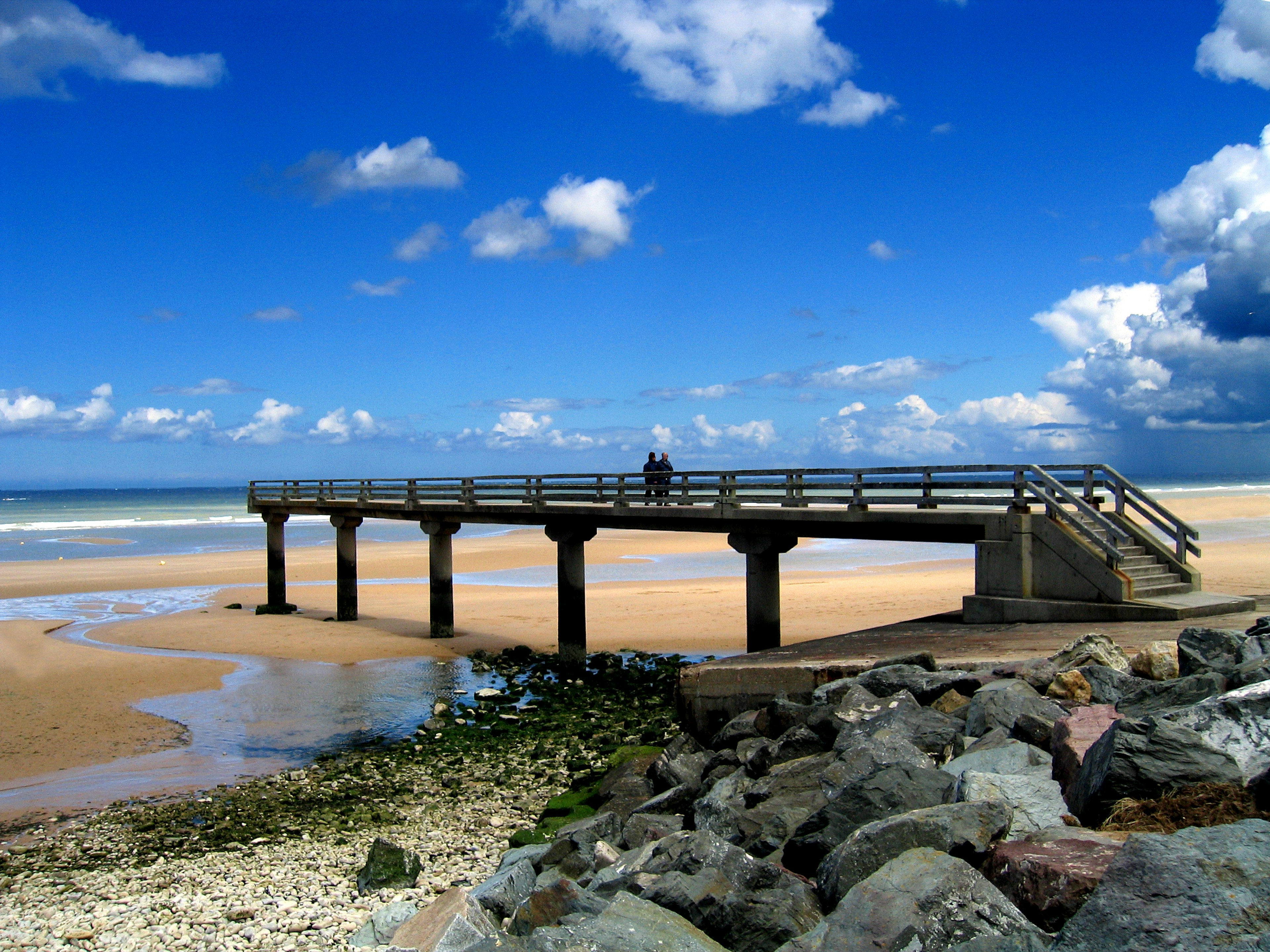 Two people standing on a pier jutting over the golden sand of Omaha Beach under bright blue skies, with the sea in the distance