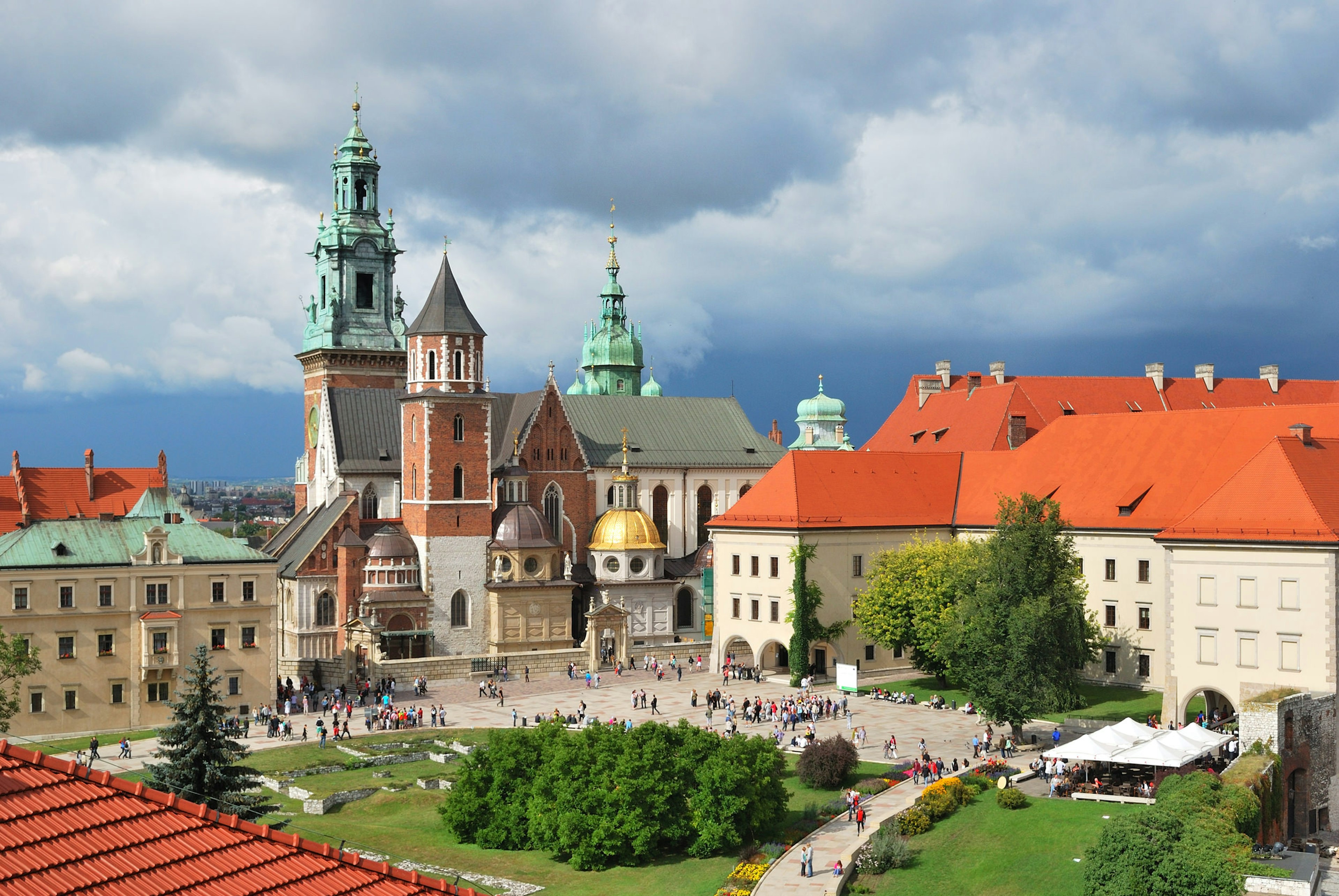 A large cathedral building in Gothic style is surrounded by other grand buildings. People can be seen moving around the grounds at the front.