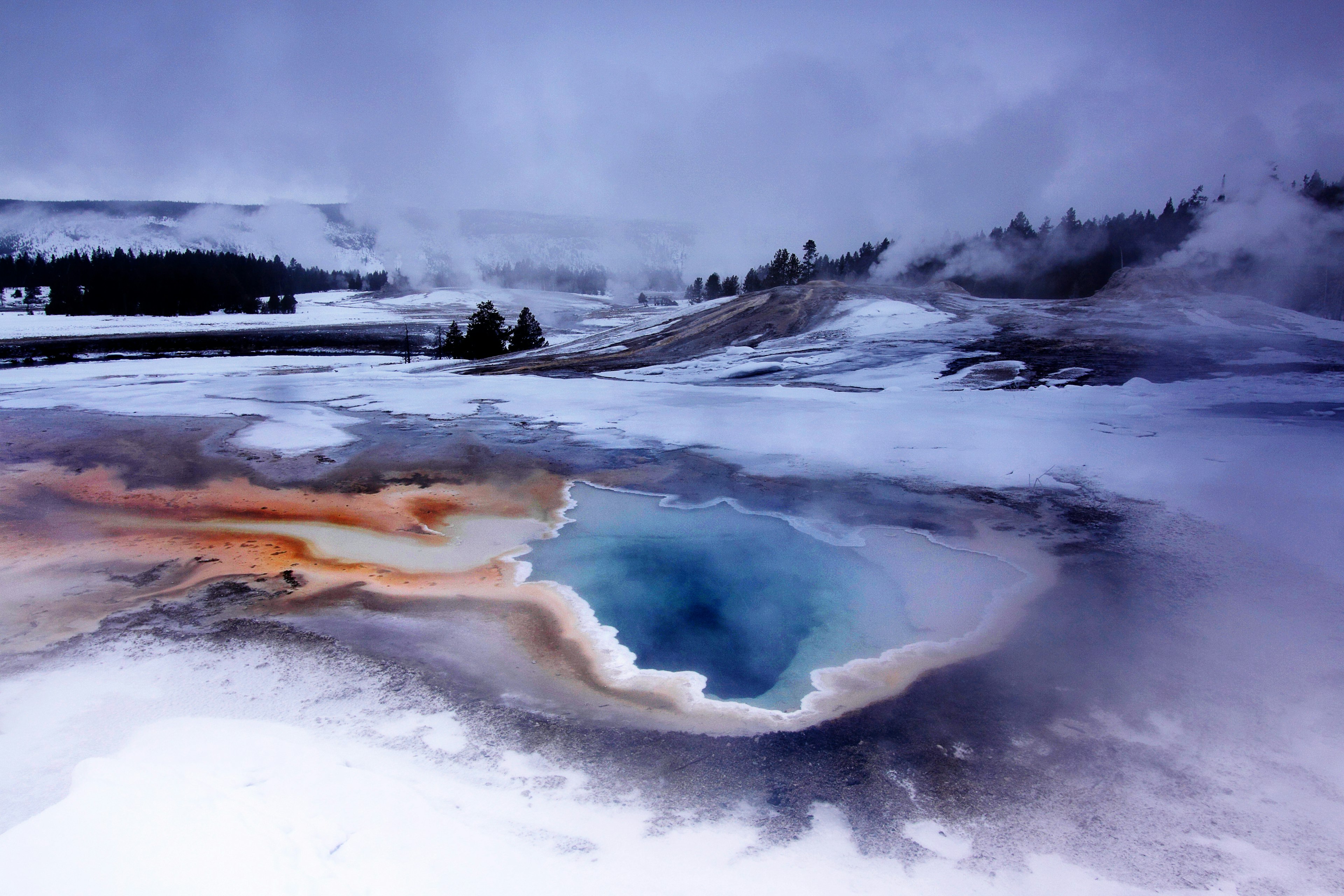 Snow and steam around a colourful water pool near Old Faithful geyser during winter.