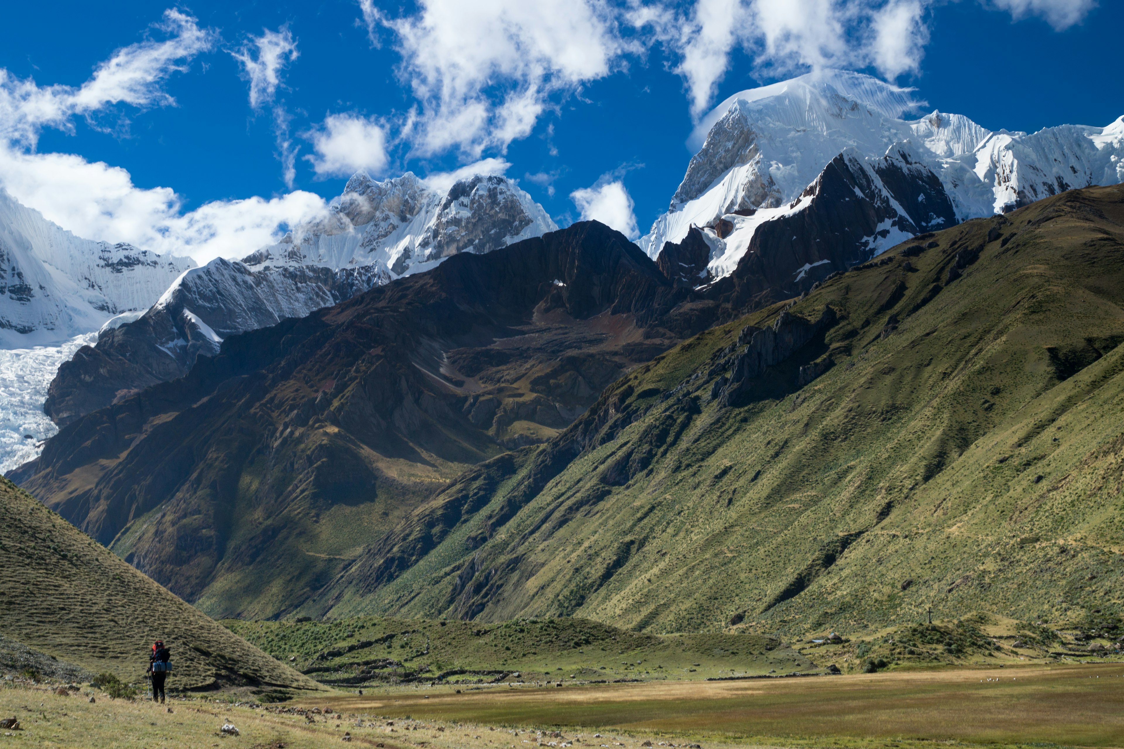 A trekker walks with green hills and rocky peaks in front of them