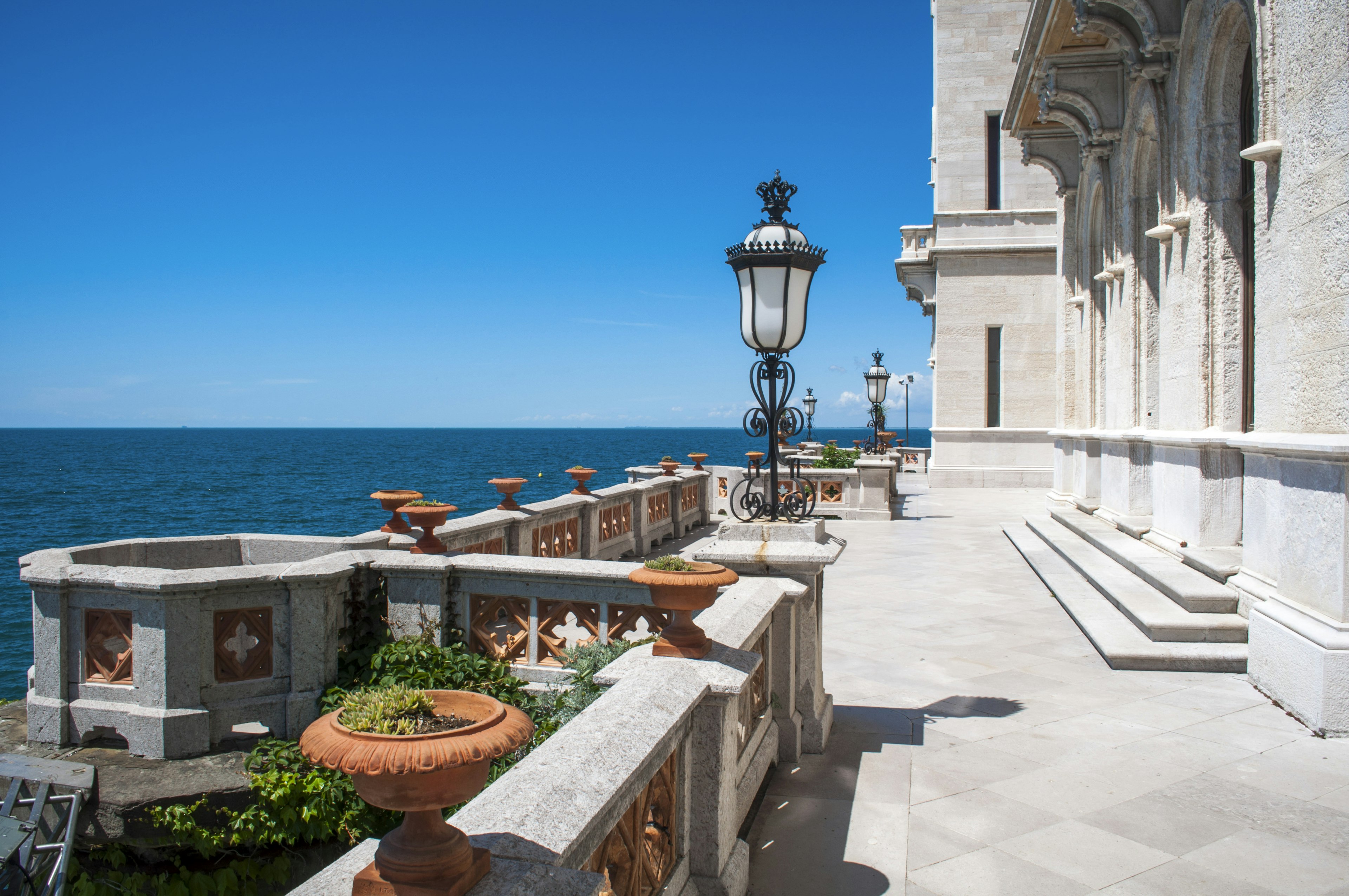 The balcony of a stone neo-Gothic castle, lined with plants in pots and wrought-iron lamps.