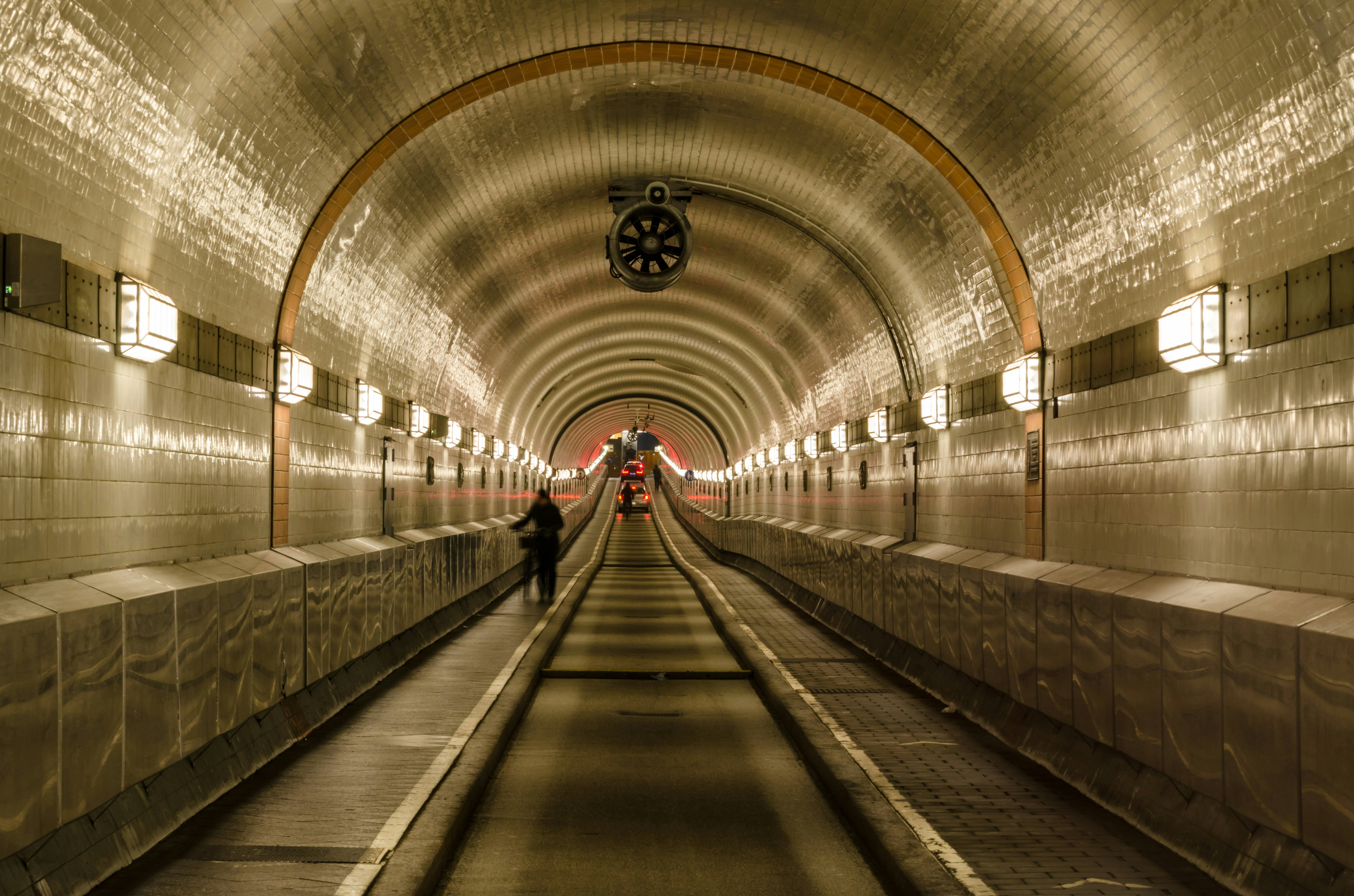 An image taken inside the Old Elbe Tunnel, Hamburg. The domed tunnel is tiled with white and black tiles and illuminated by old-fashioned lights every few metres. someone is wheeling their bike through the tunnel and in the far distance we can see red brake lights.