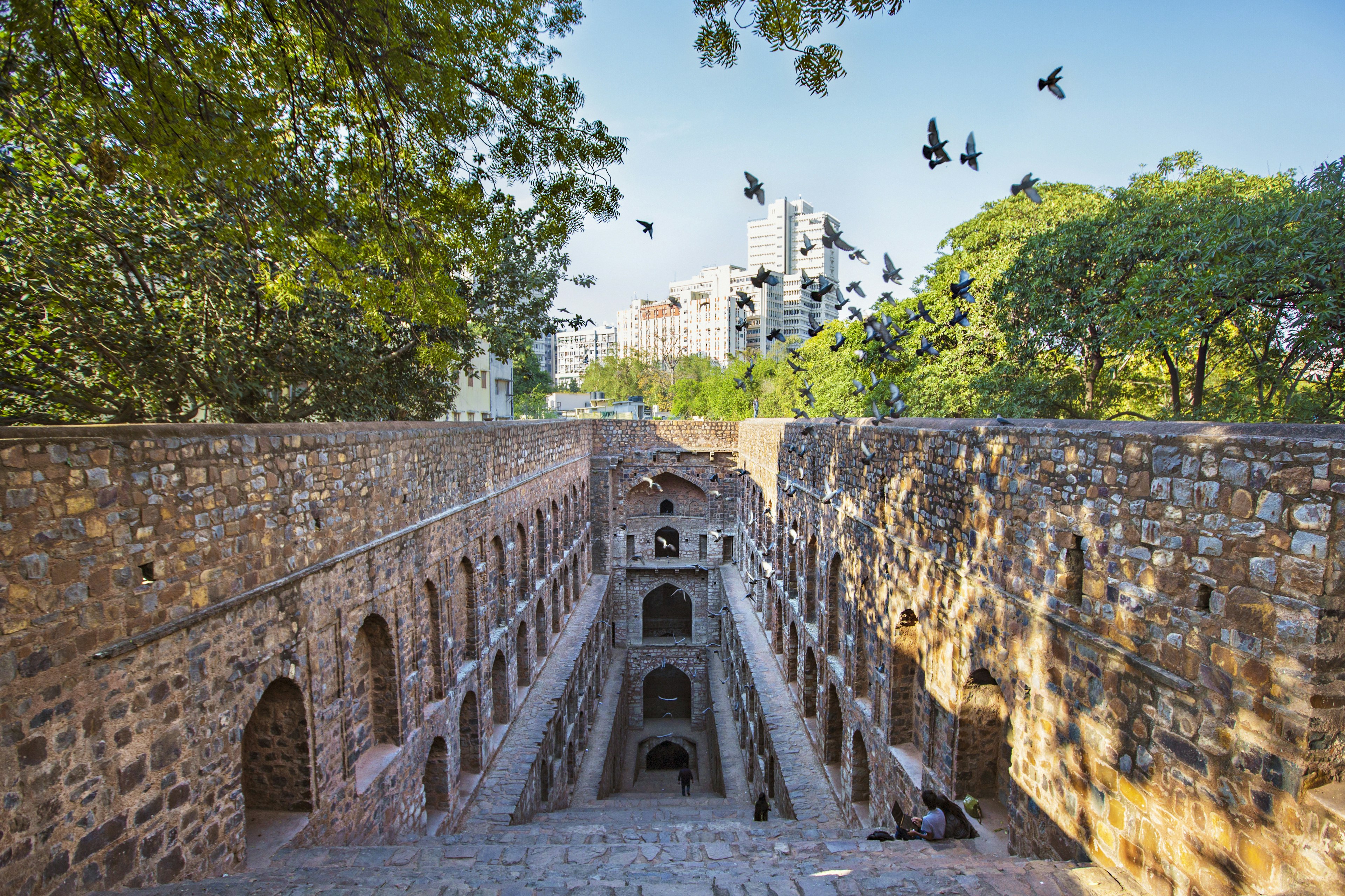 The stone Agrasen Ki Baoli stepwell in Delhi. A staircase descends downwards, enclosed by a vertical wall on all other sides, which are adorned with shallow arches. A few people sit on the steps, while, back on ground level, some trees and buildings are visible around the stepwell.