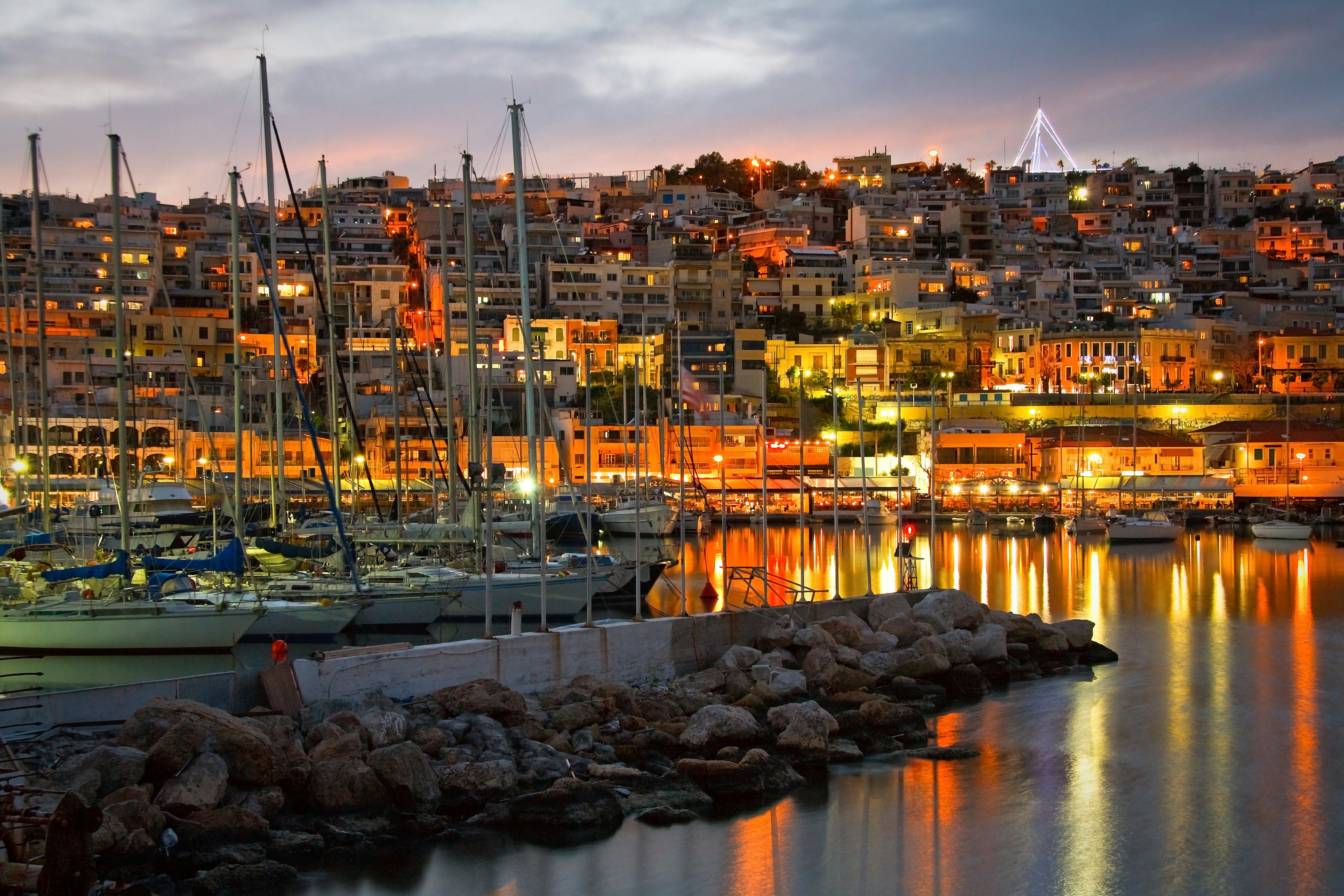 A view looking up at a hillside in Piraeus covered with buildings; the sun is setting and the streetlights are one, giving everything an orange glow. Piraeus, Greece.