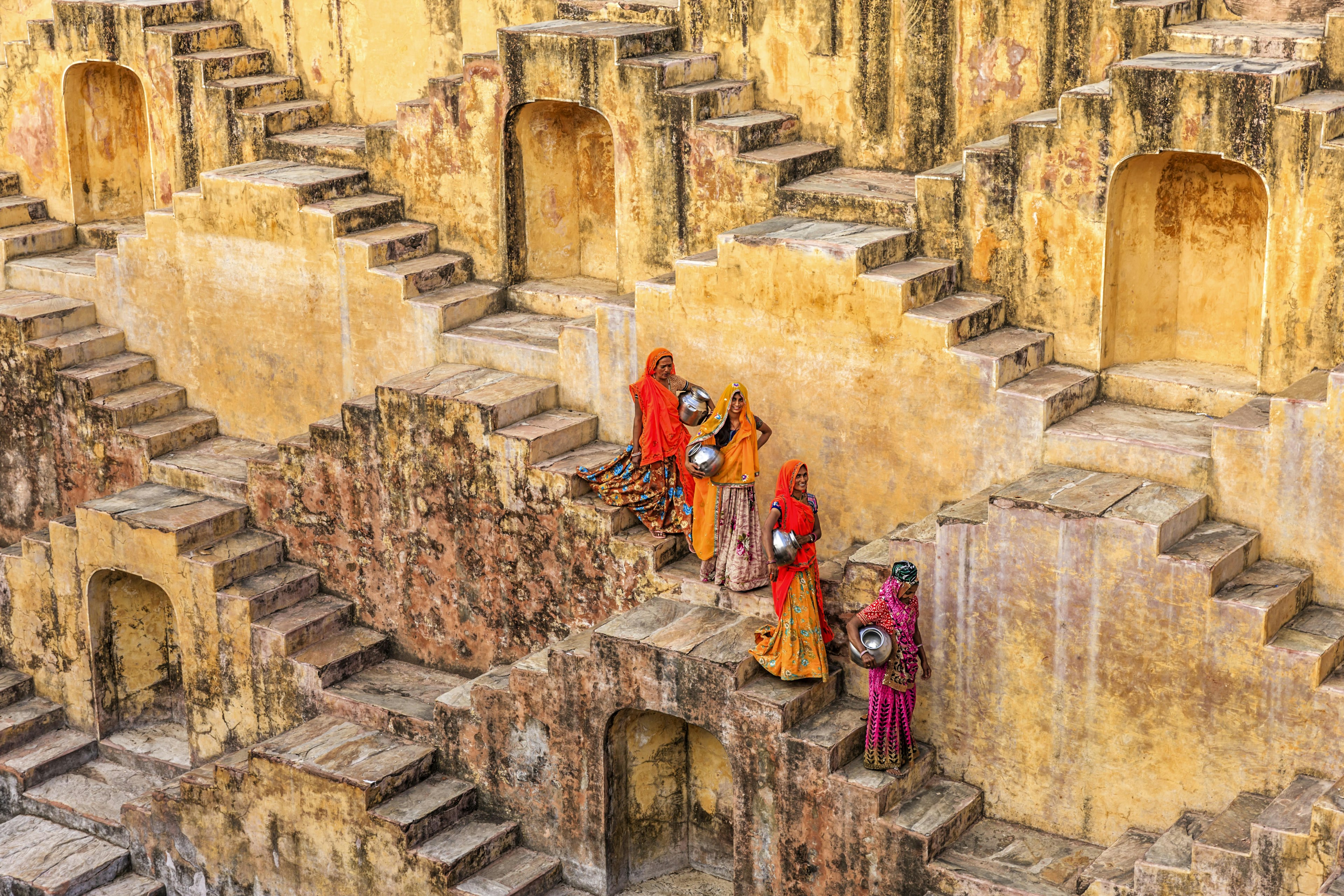 A posed picture of four women, carrying metallic pots, walking down the stairs of a stone stepwell in Rajasthan, India. The four women wear colourful saris that stand out against the stone background.