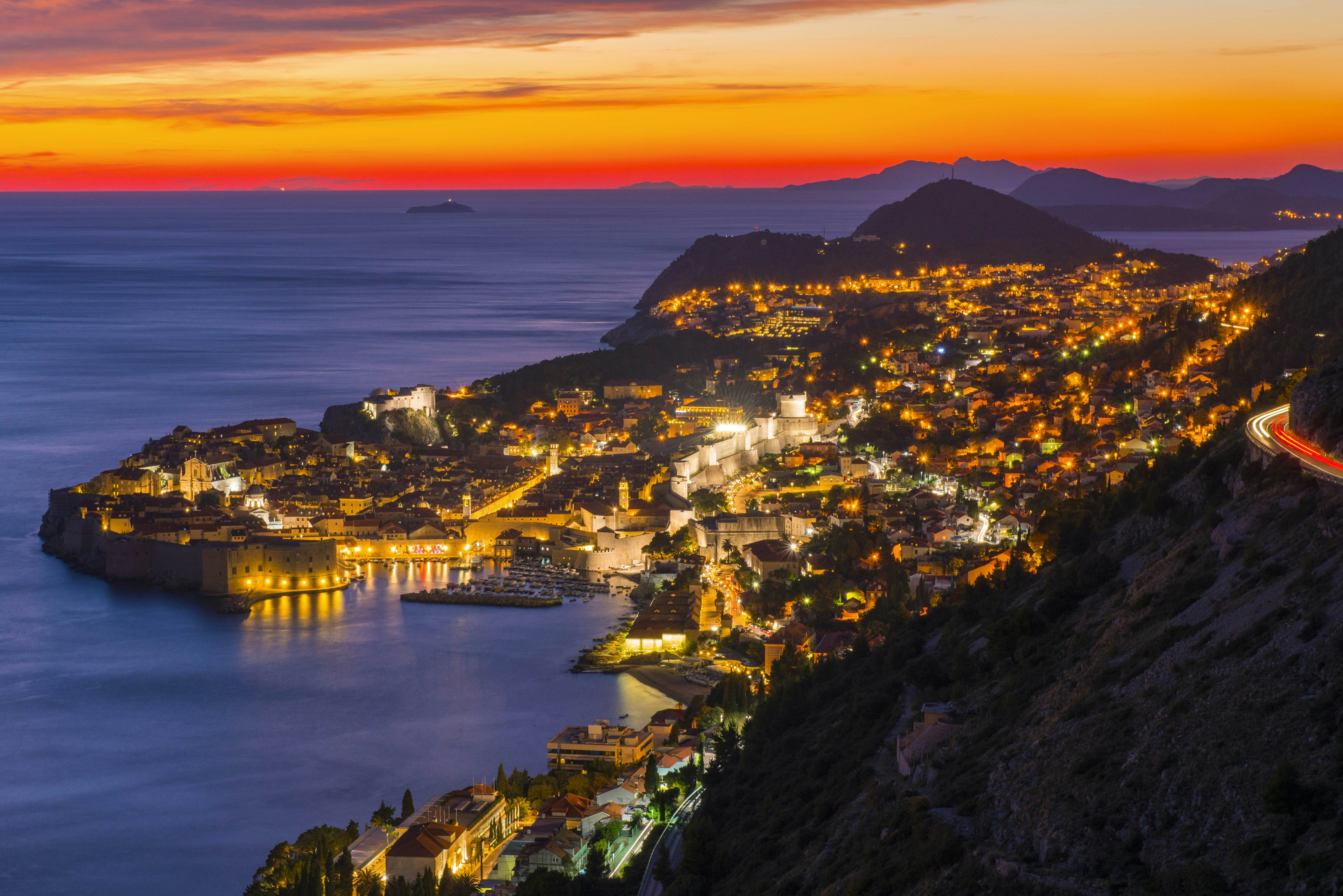 The sky glows bright orange against a navy ocean as the sunset casts a glow over a seaside city with medieval walls