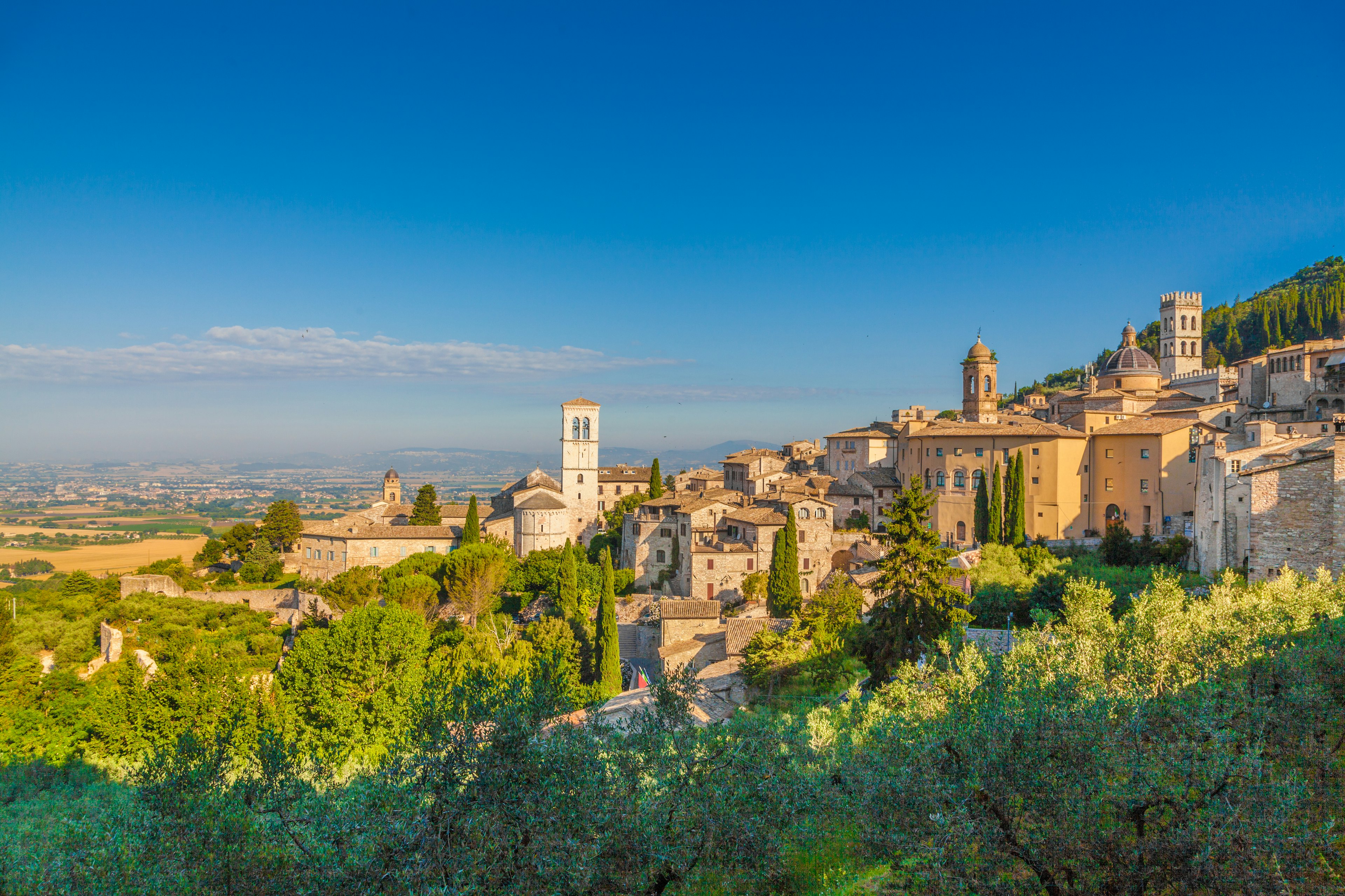 The sandstone-coloured buildings of Assisi glow in the sunshine, with a clear blue sky behind and countryside in the distance