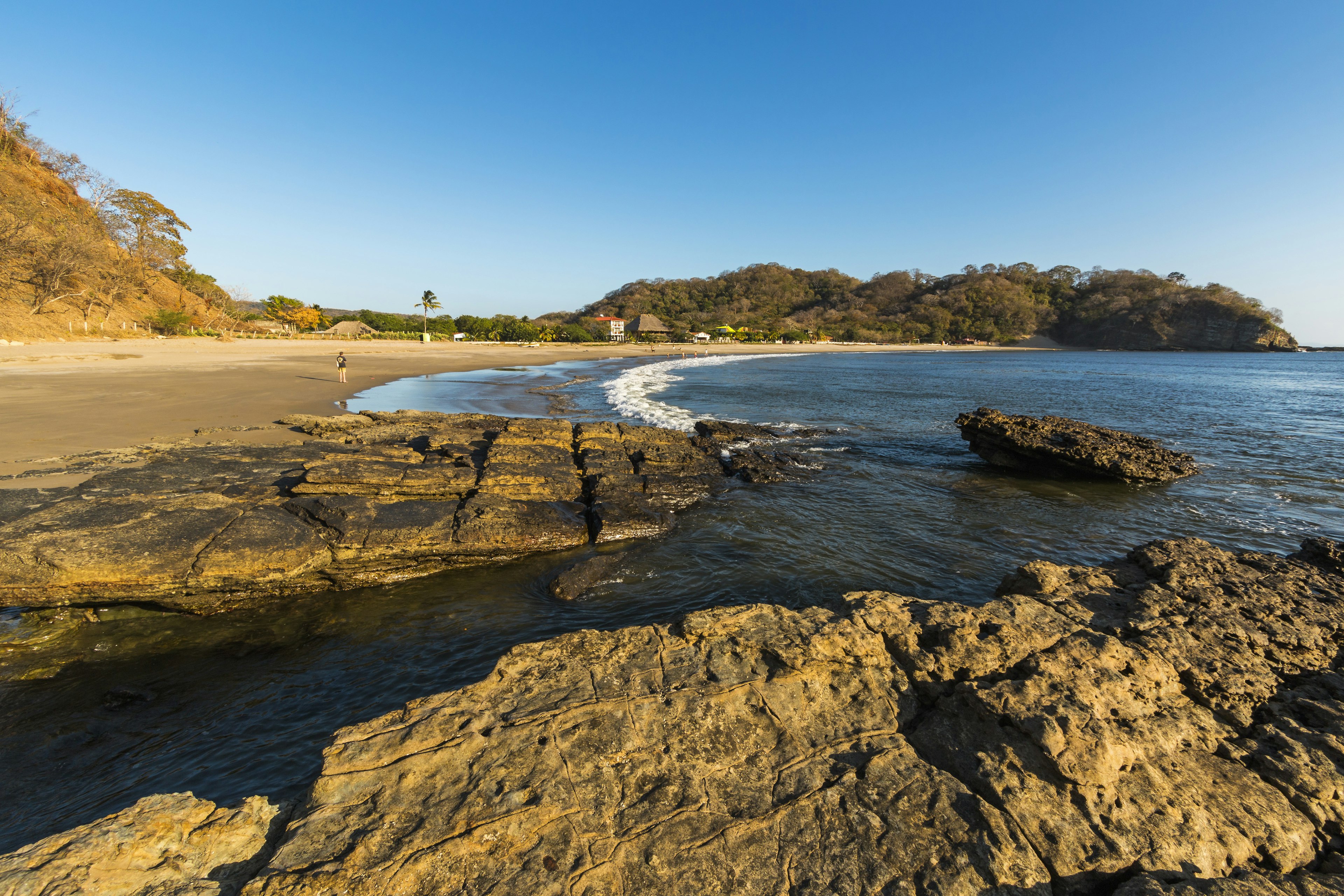 A person stands on a beach looking out on the sea as waves crash on the surf. There are jagged rocks jutting into the sea; best beaches Nicaragua