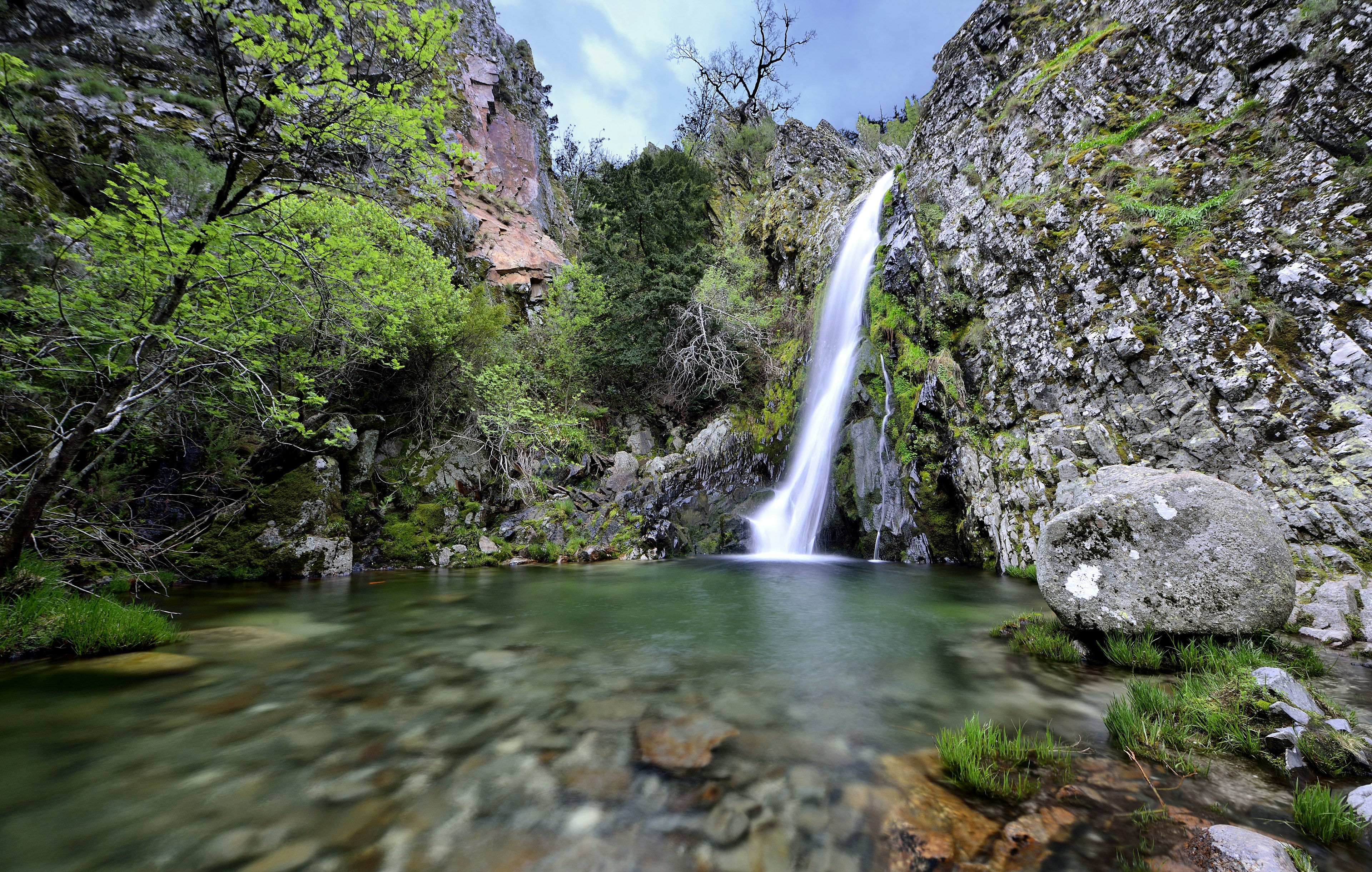 Long exposure image of a waterfall and lake, dense foliage and large rocks throughout.