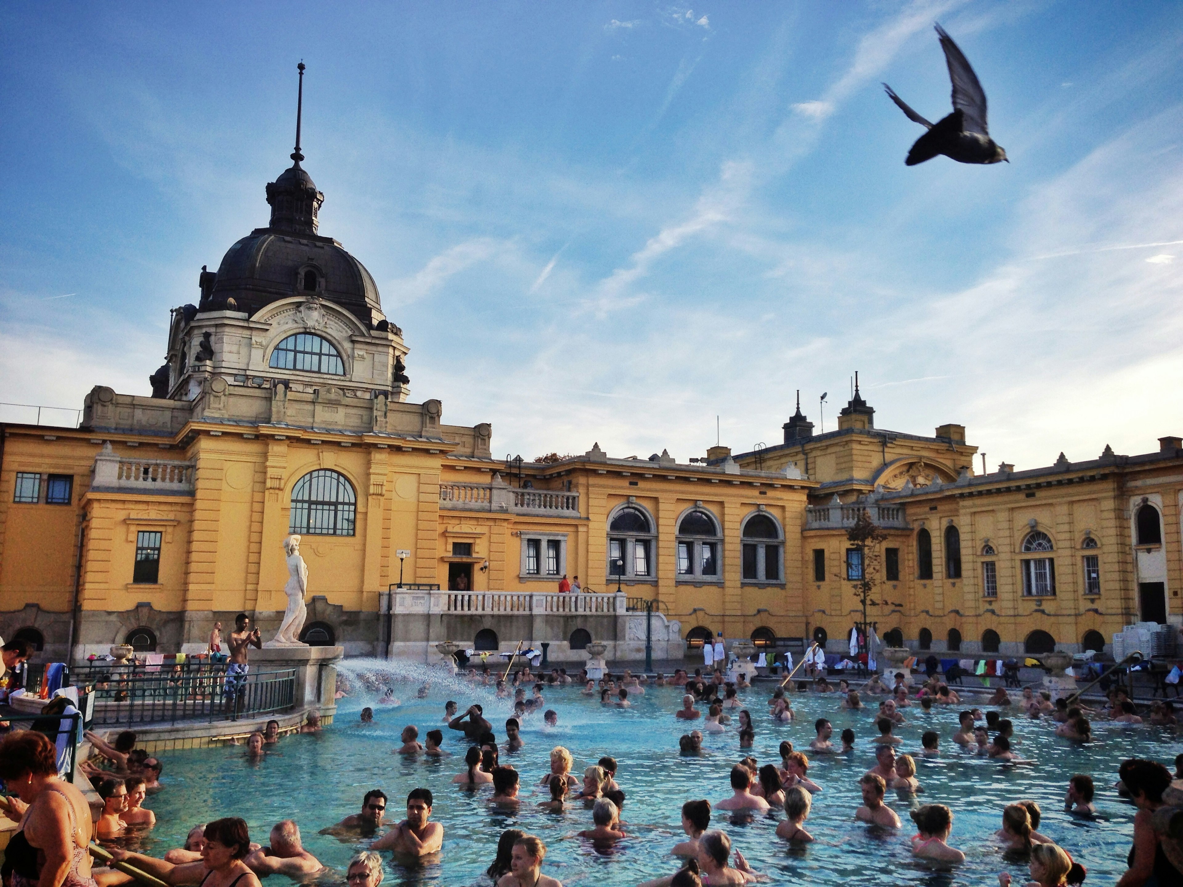 A clear blue swimming pool with an elaborate yellow building in the background, filled with many swimmers.