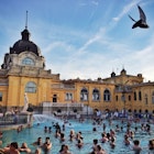A clear blue swimming pool with an elaborate yellow building in the background, filled with many swimmers.
