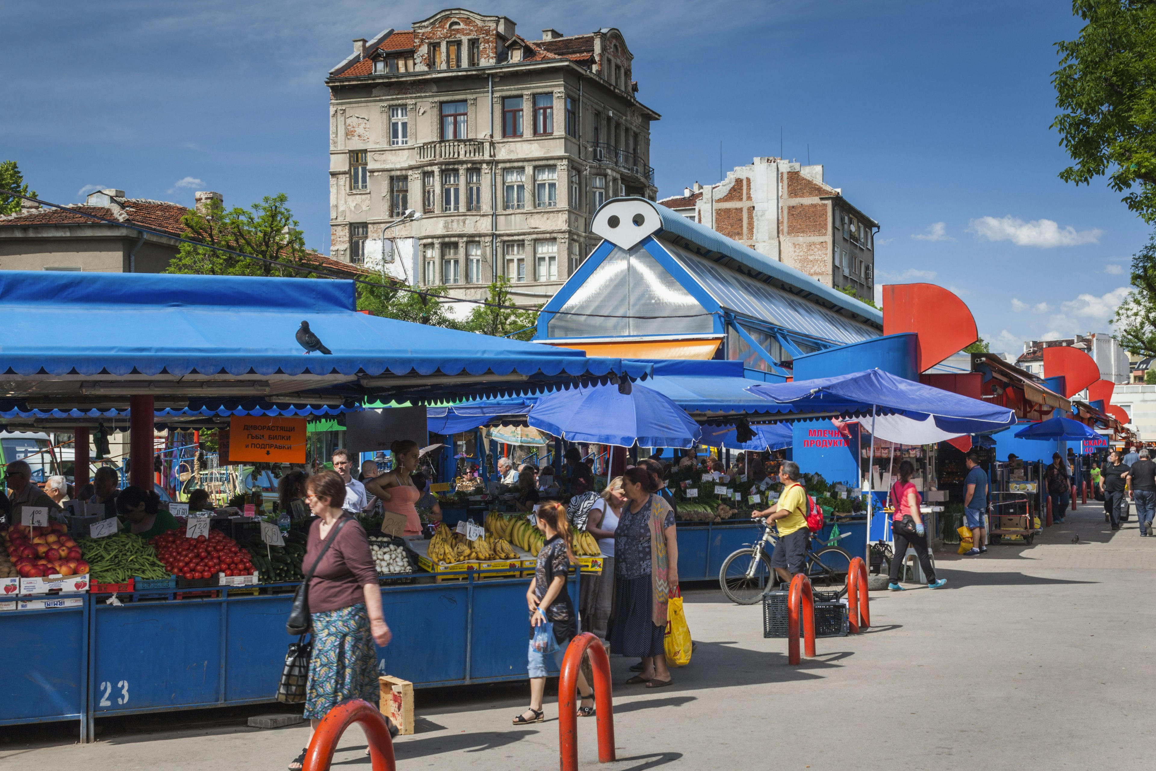 Shoppers stroll by a series of stalls packed with fruits and vegetables under a bright blue canopy.