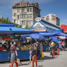 Shoppers stroll by a series of stalls packed with fruits and vegetables under a bright blue canopy.