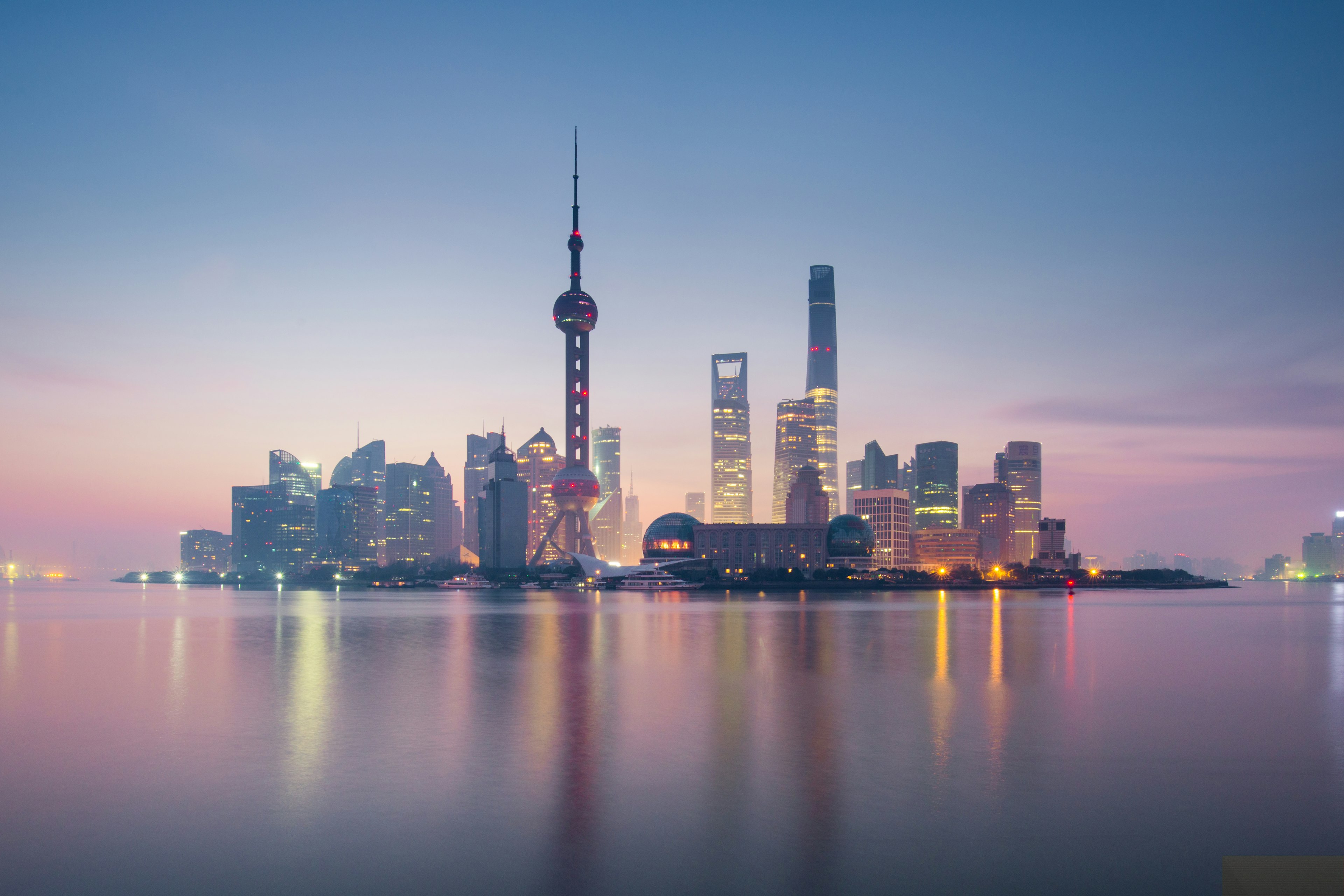 The skyline of Pudong in Shanghai, surrounded by pink water and pale pink and blue light in the sky as the sun sets