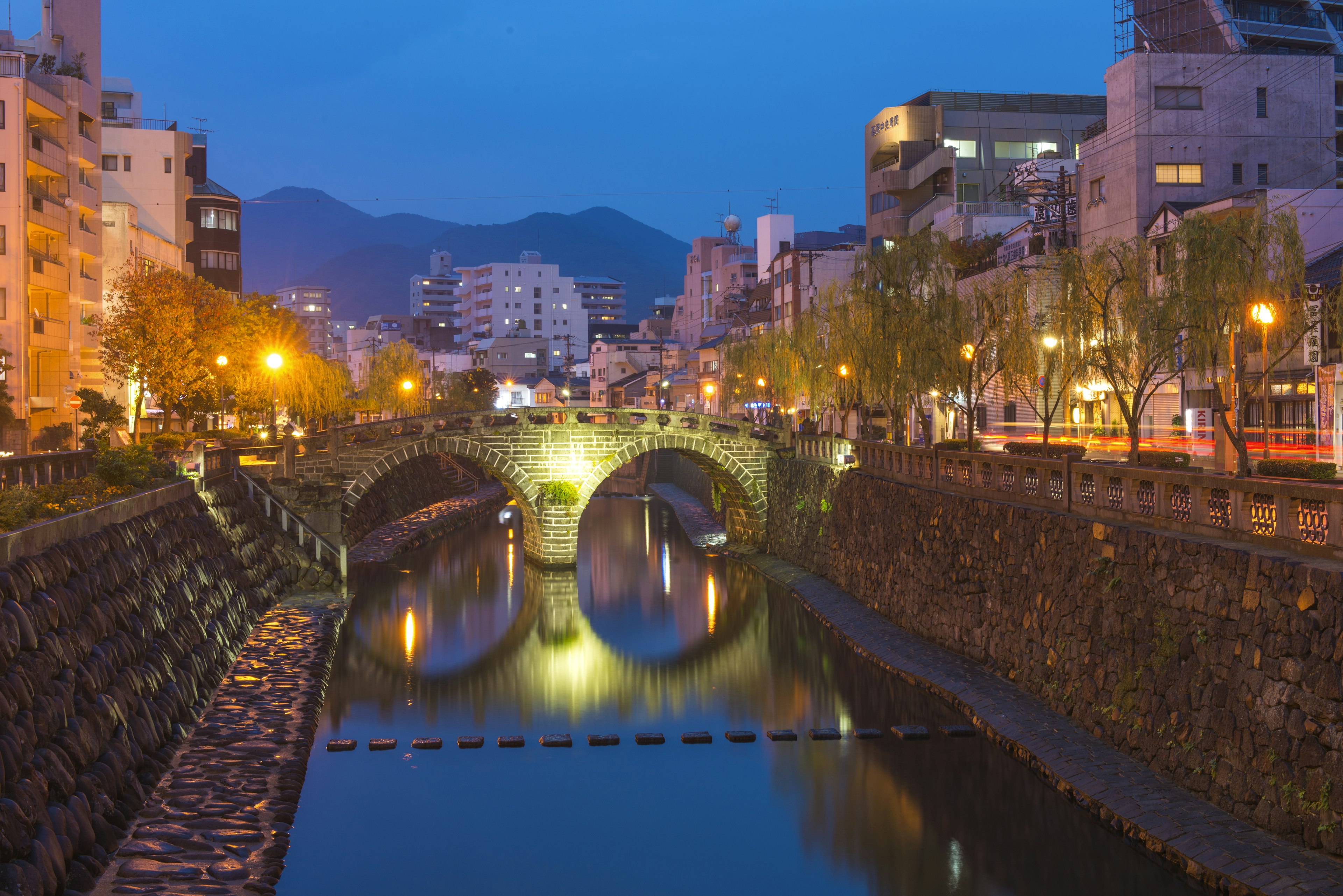 A bridge with two arches, reflected in the water, spans a steep canal in Nagasaki.