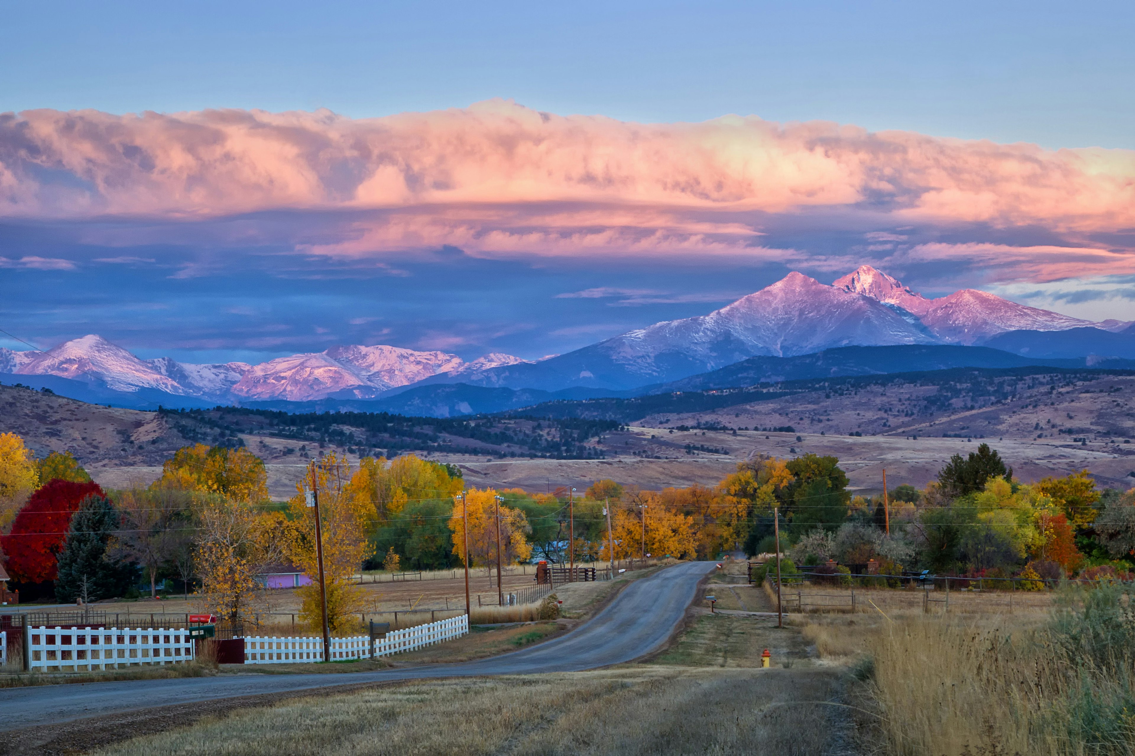 Long's Peak lights up at sunrise as a rural country road leads into the fall trees.