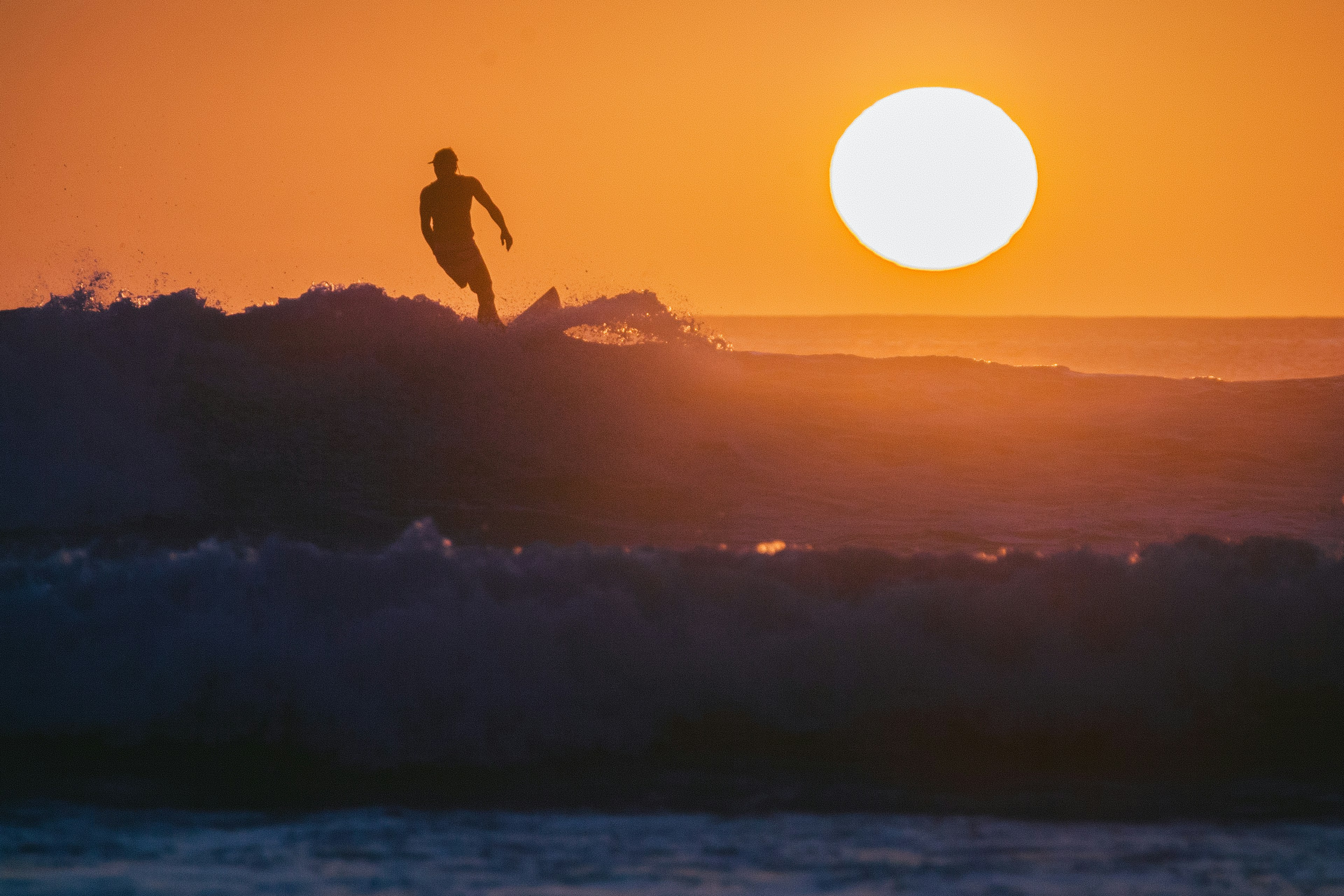 The silhouette of a surfer riding a wave with the setting sun over the ocean.