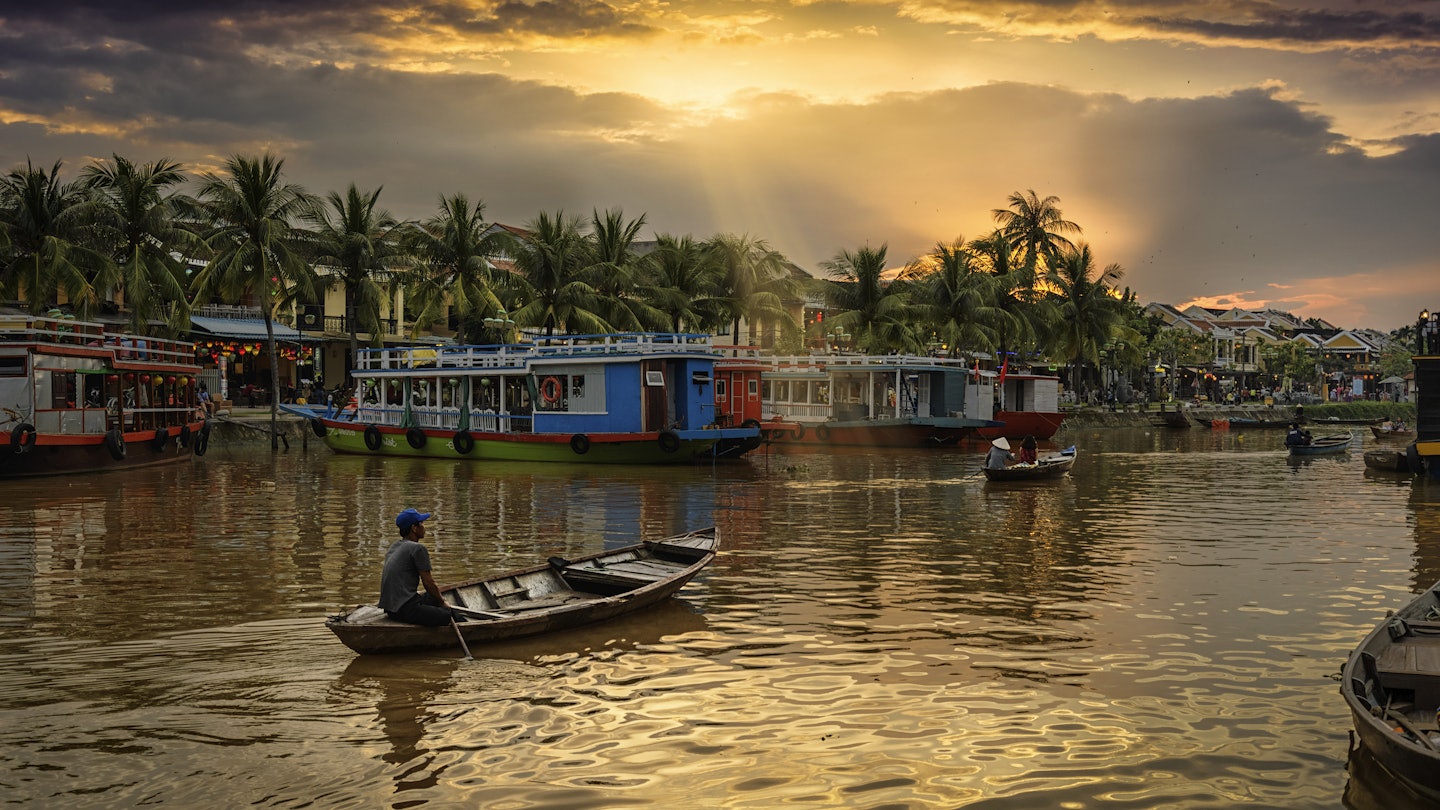 A few small wooden boats on the Thu Bon river with the sun setting over palm trees. There are big, colourful boats docked along the banks and restaurants beyond them.