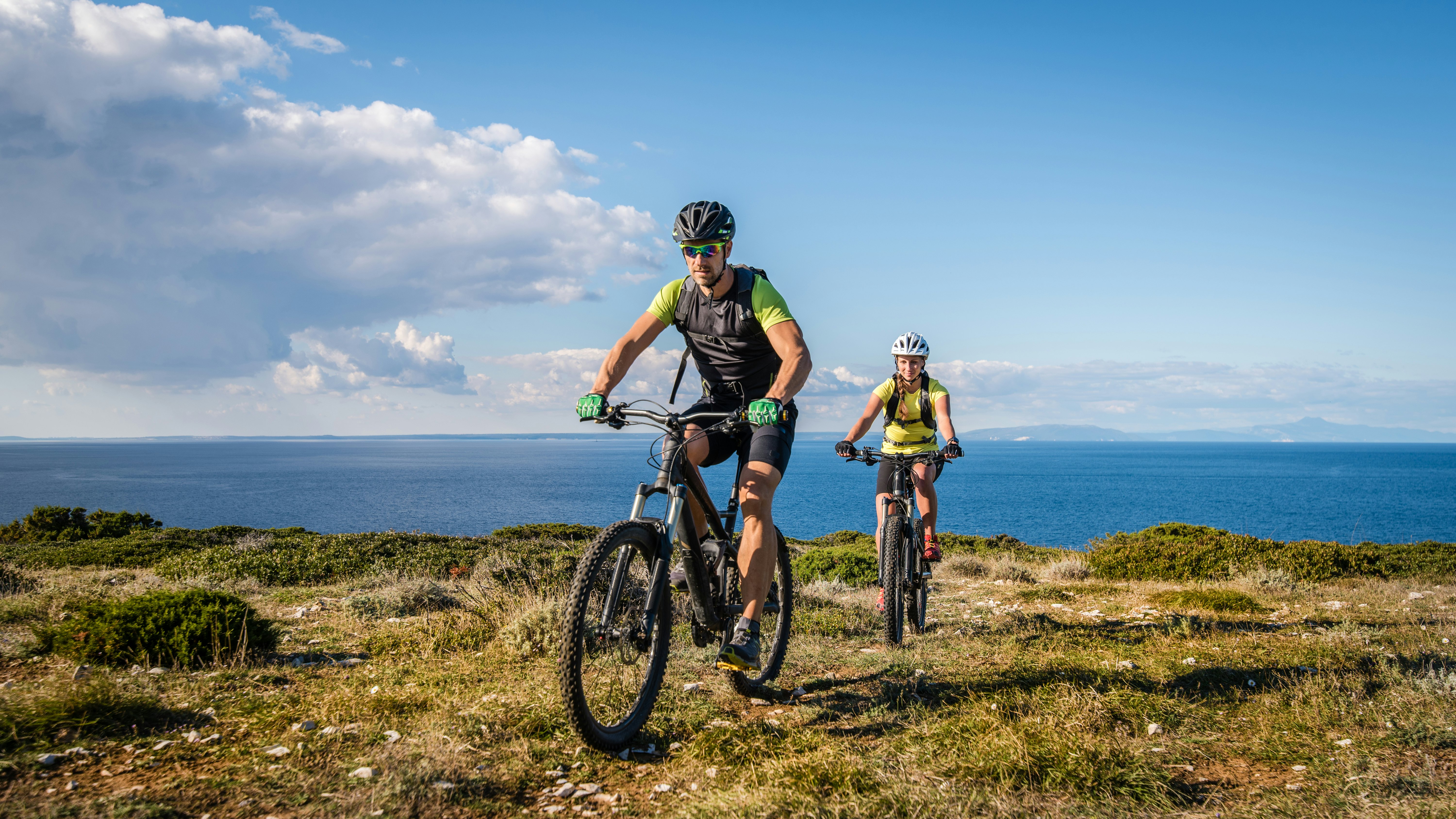 A few EuroVelo routes take in Croatia: here we see a young couple on mountain bikes cycling along a grassy headland, with the blue sea beyond.