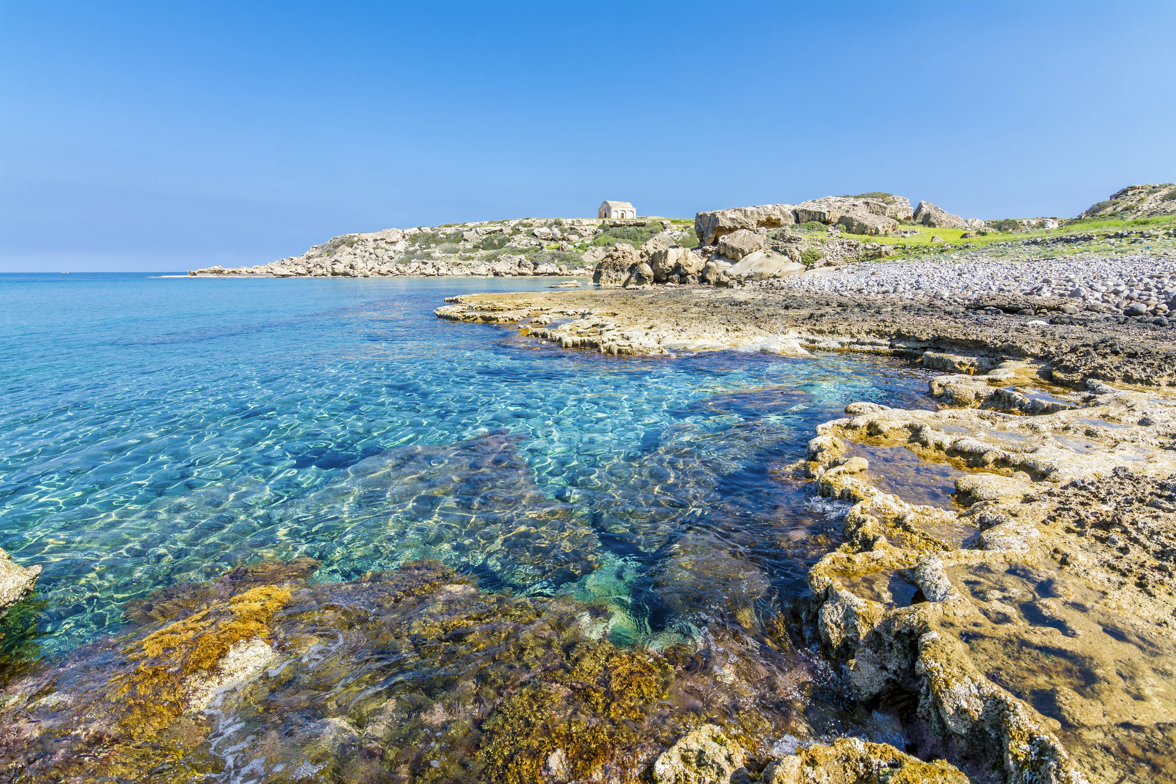 A rocky shoreline with a small stone building in the distance. The sea is clear and very still.