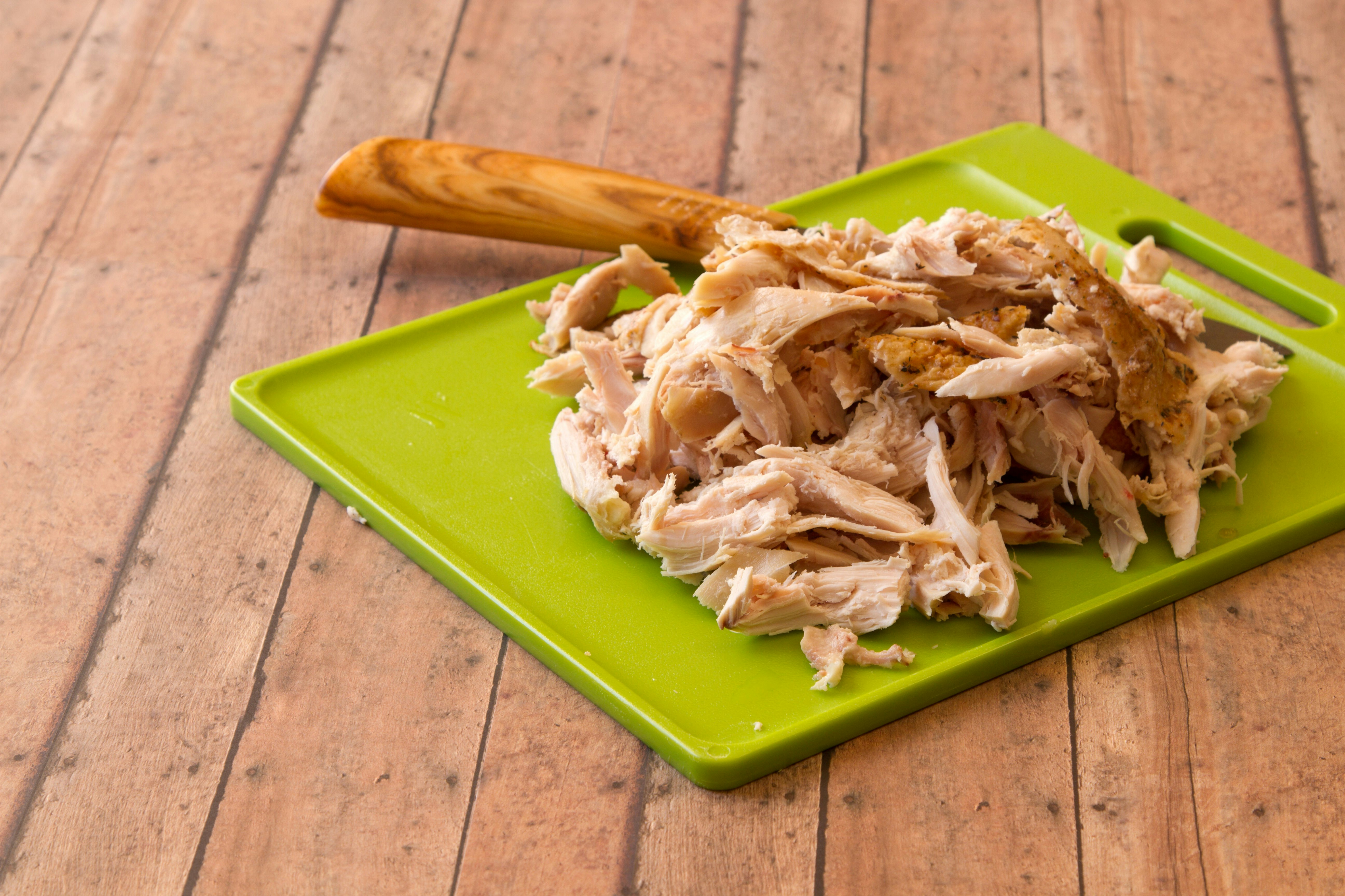 Shredded rotisserie chicken on a green plastic cutting board and carving knife against wood plank background